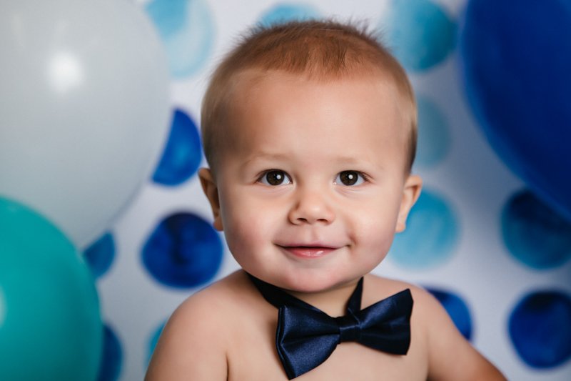 A young boy with a shaved head and wearing a navy blue bow tie, smiling at the camera, surrounded by blue and white balloons.
