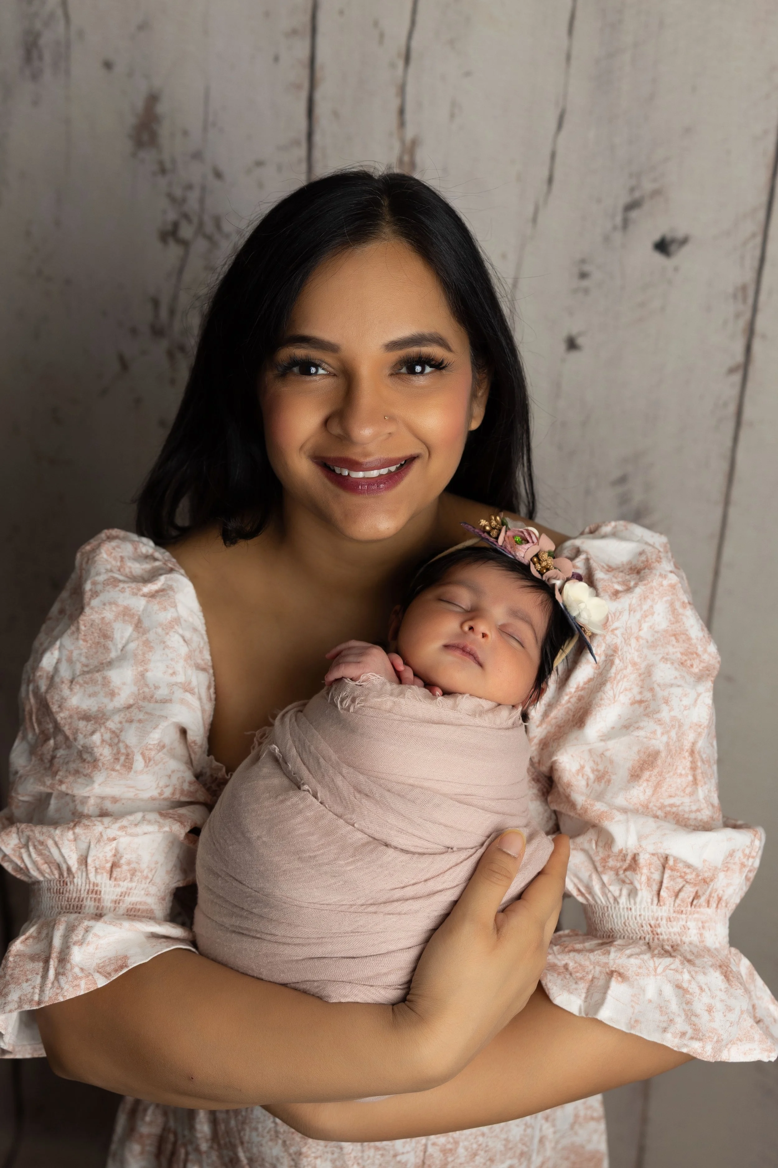 A smiling woman holding a sleeping baby girl wrapped in a pink cloth, with a floral headband, in front of a wooden background.