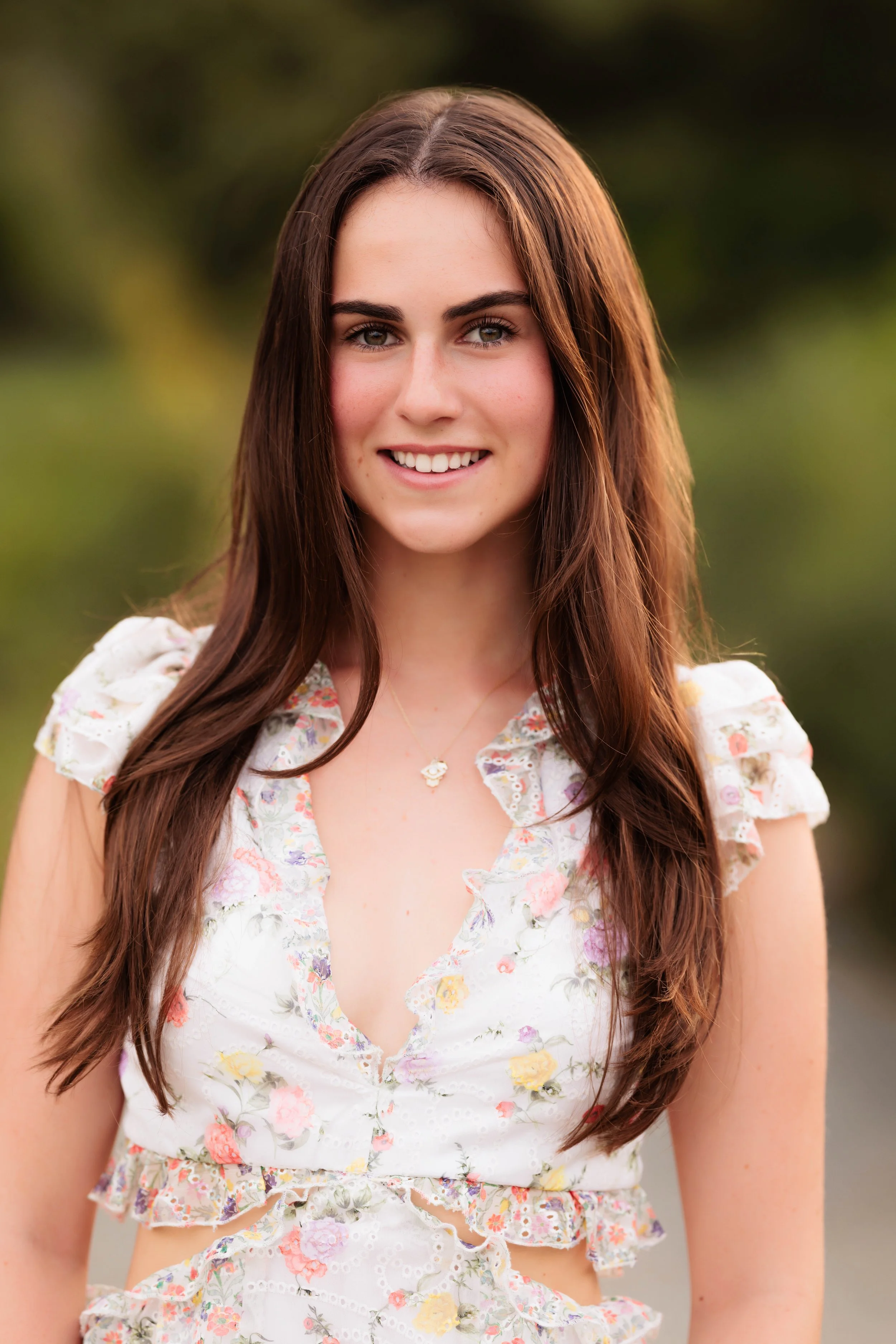 A young woman with long brown hair and blue eyes smiling outdoors, wearing a floral crop top with ruffled sleeves and a necklace.