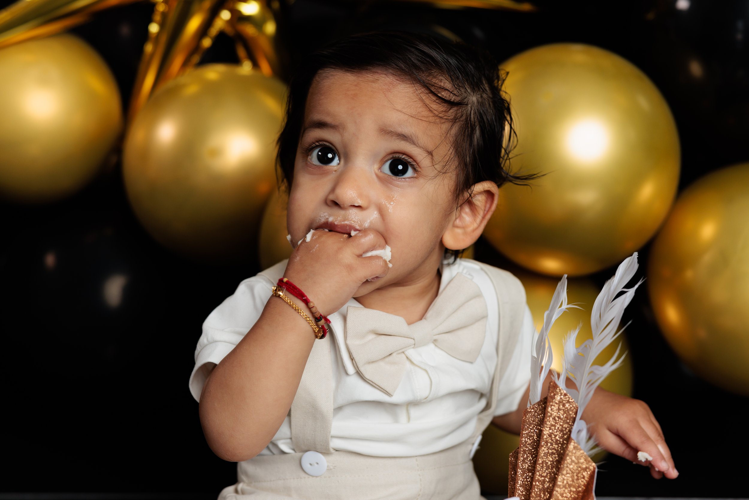 A young child with dark hair, wearing a cream-colored shirt and bow tie, is eating cake with a background of gold balloons.