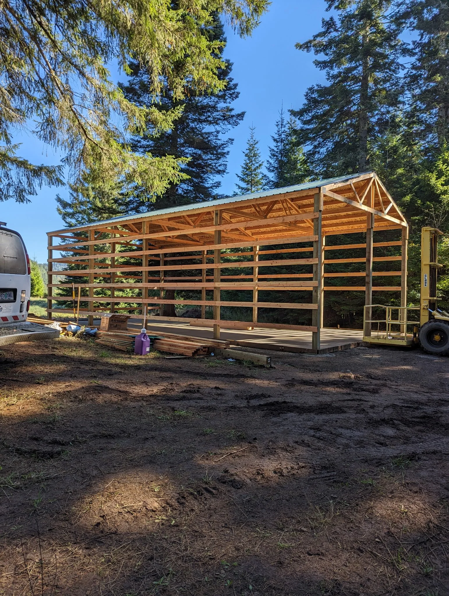 A wooden structure under construction in a forested area, with tall pine trees and a clear blue sky in the background.