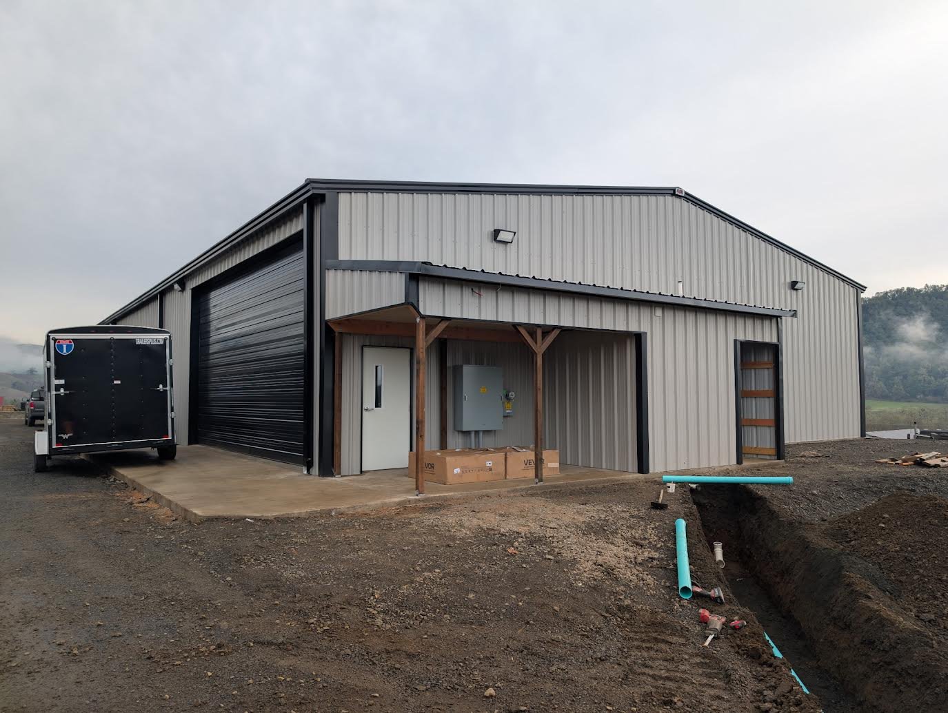 A newly constructed metal building with a large roll-up garage door, a side door, and a small covered porch area, with construction materials and pipes nearby.