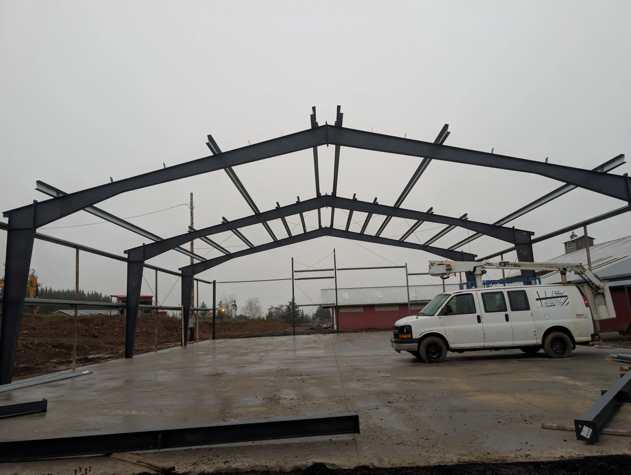 Construction site with steel framework and a white utility truck parked on a concrete ground under an overcast sky.