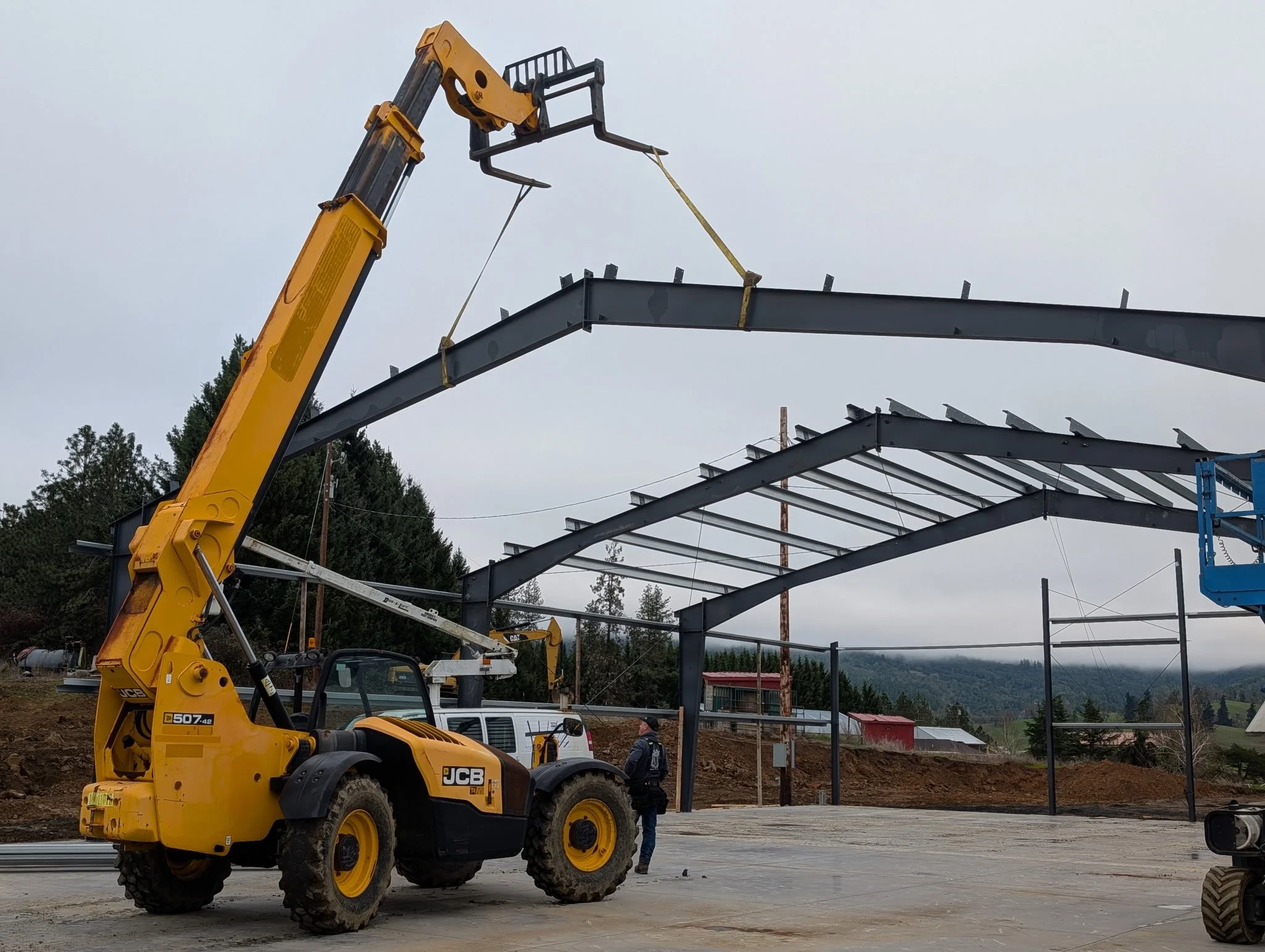 Construction site with a yellow JCB backhoe loader and steel framework for building structure. Sky is overcast, and trees and distant buildings are visible in the background.