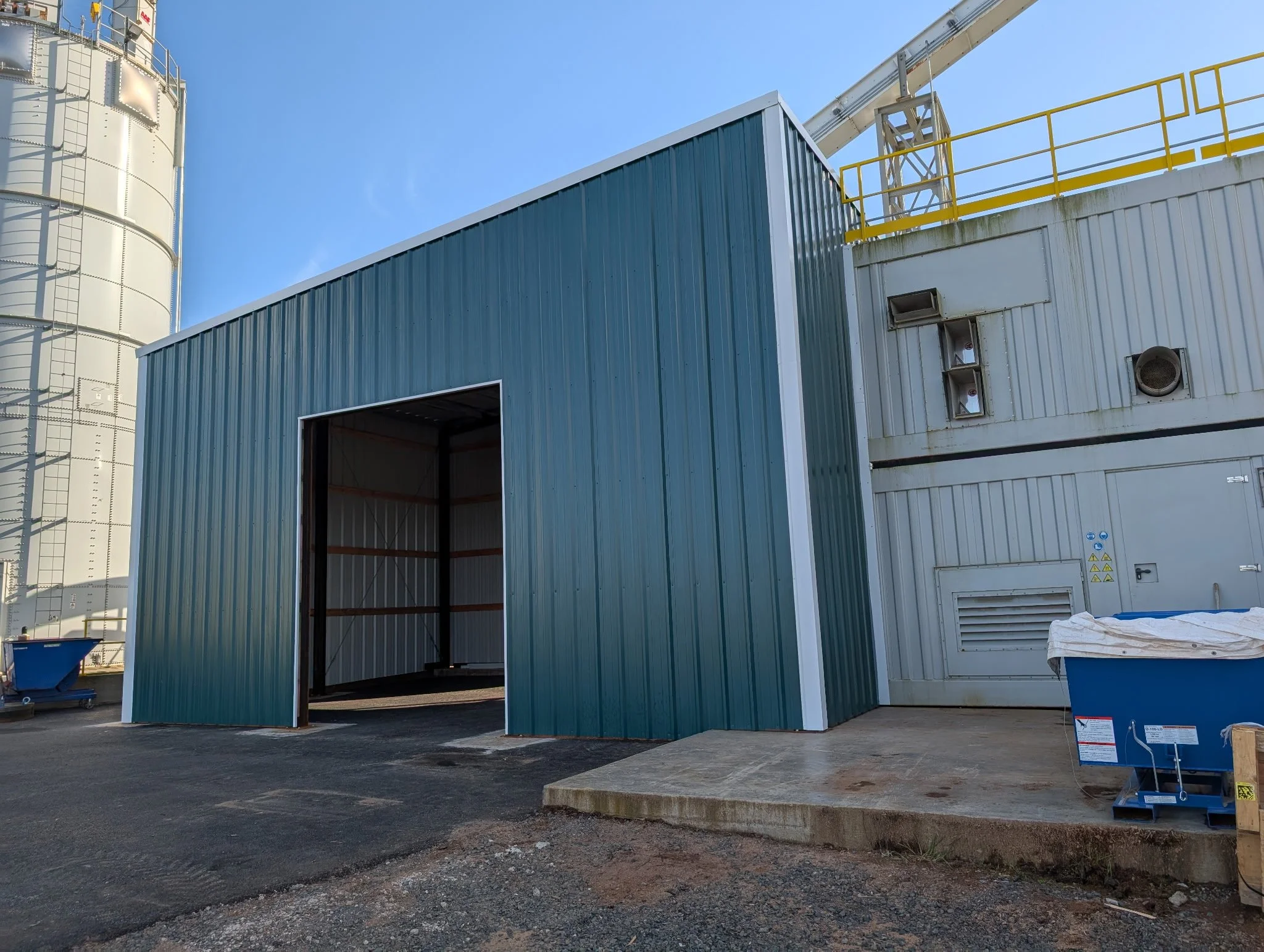 A metal industrial building with an open garage door, with a large silo to the left and a crane above. There is a table with supplies and warning signs on the building wall.