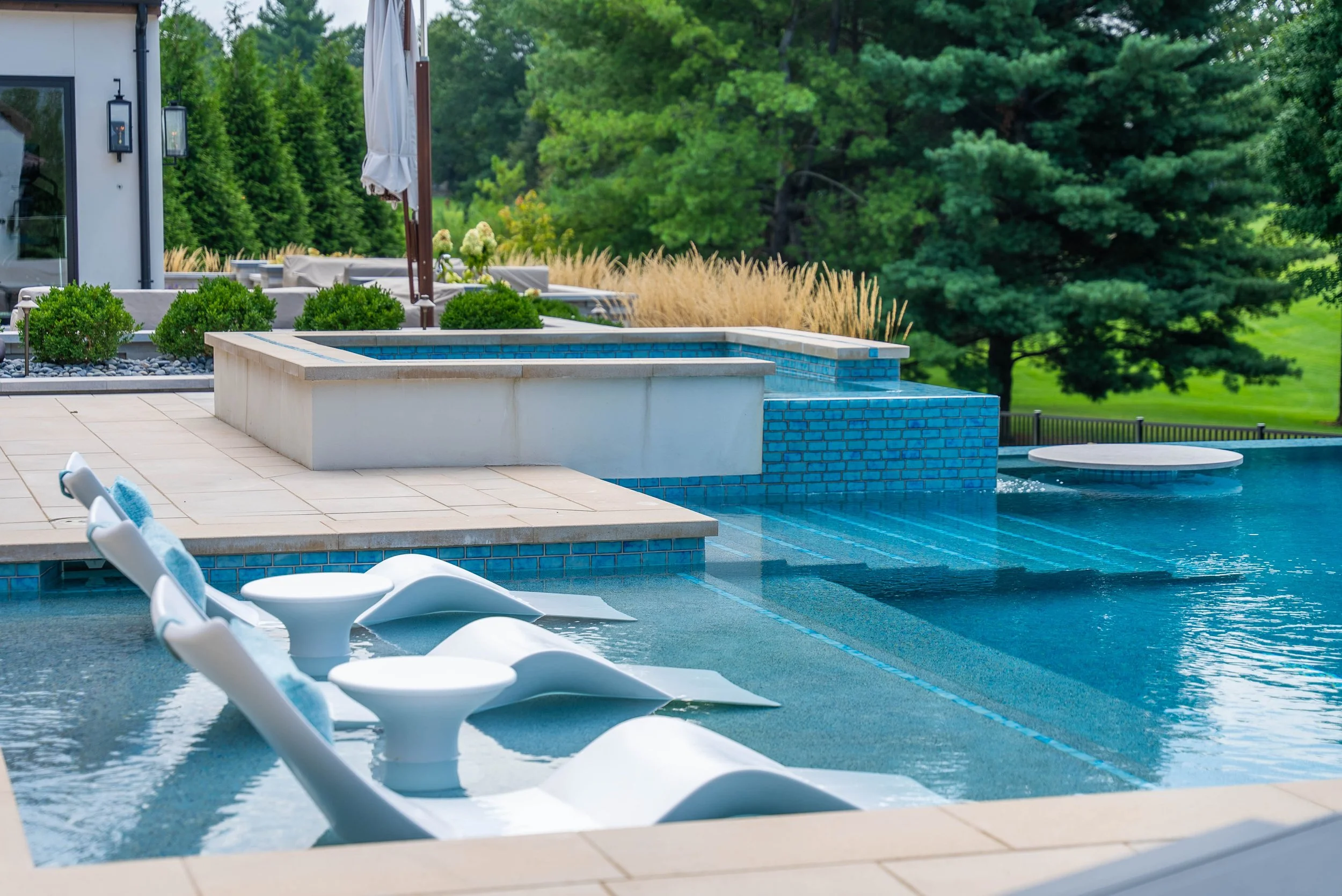 Luxury backyard swimming pool with built-in seating, water feature, and outdoor furniture, surrounded by lush greenery and trees.