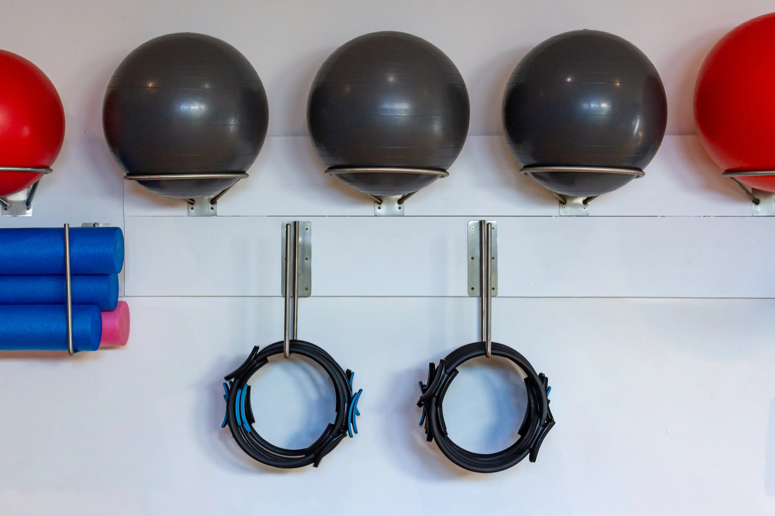 Wall with four black exercise balls mounted on wall, two resistance bands hanging below, and a piece of blue and pink foam rollers on the left.