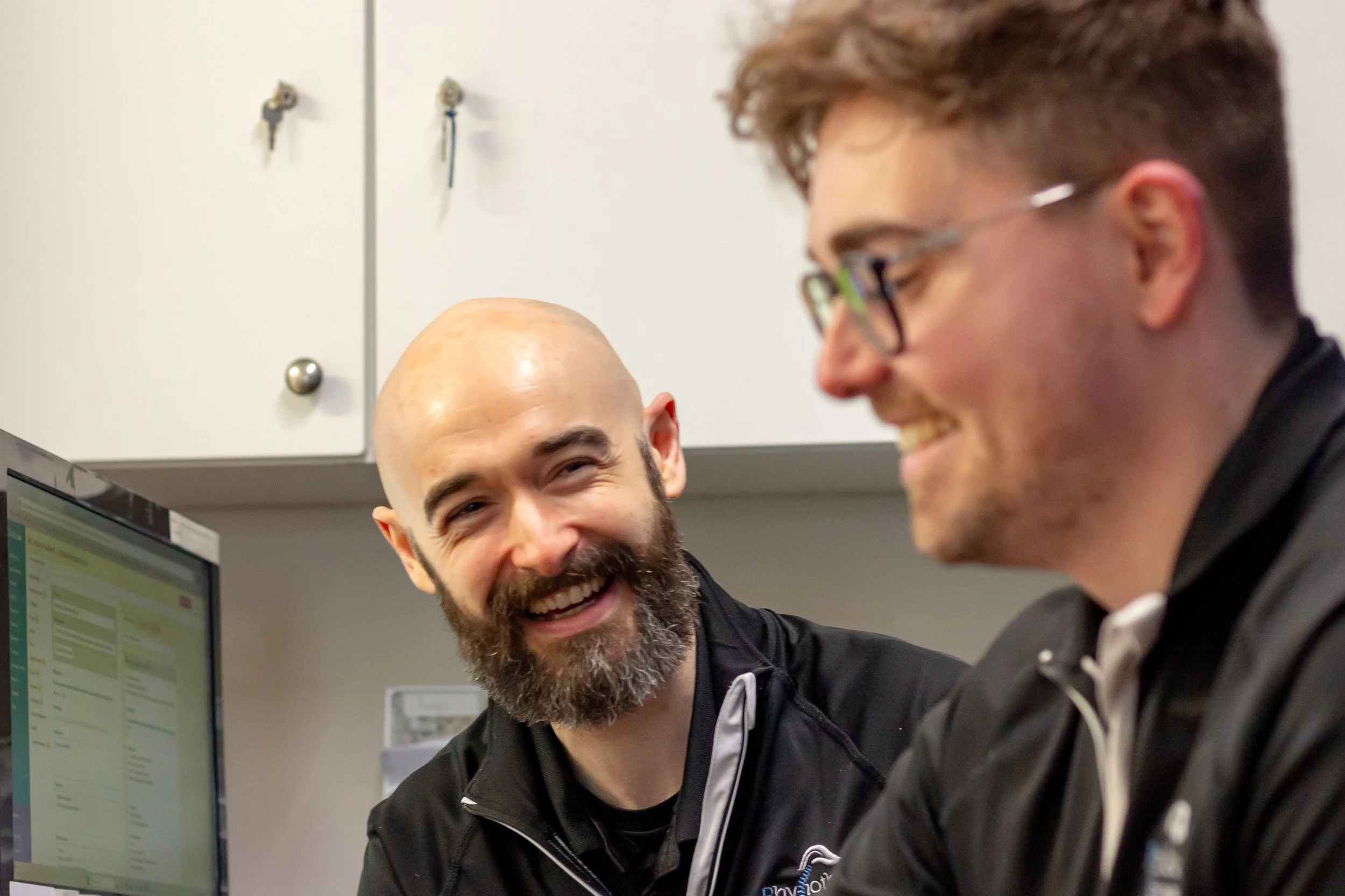 Two men working together at a computer, smiling and engaging in conversation, in an office setting.