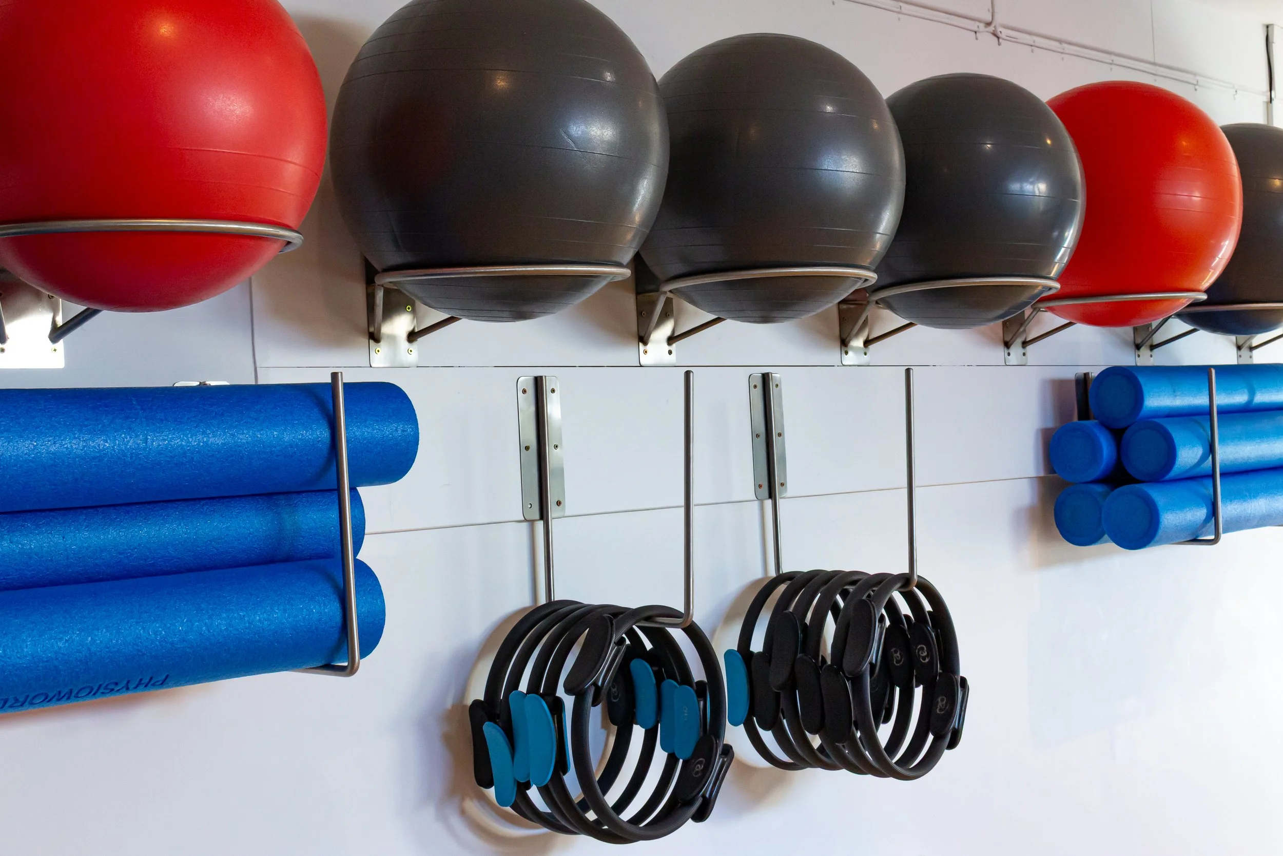 Shake weights, exercise balls, foam rollers, and resistance bands hanging on white wall in a gym.