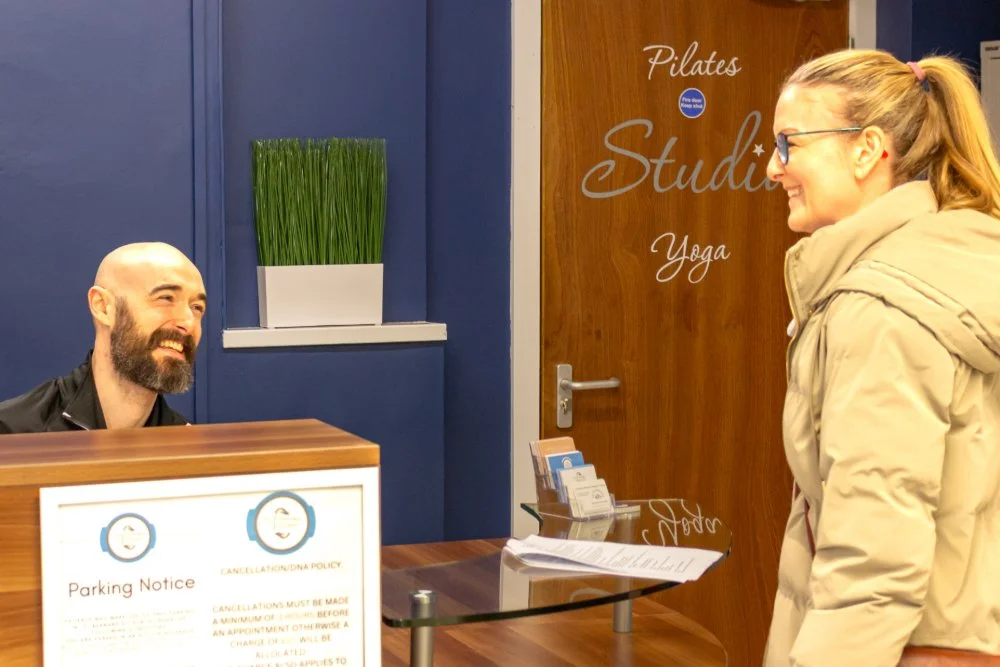 A woman in a beige jacket and glasses smiling and talking to a cheerful man with a beard behind a reception desk. The background has a blue wall, a pot with green grass, and a door with signs for Pilates, Studio, and Yoga.