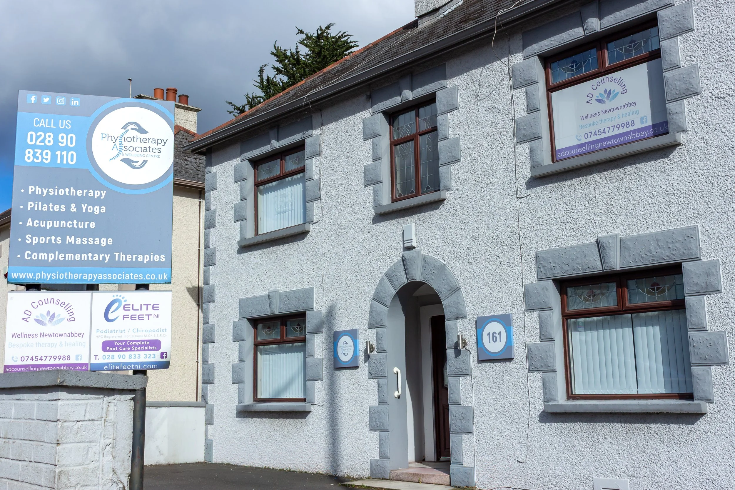 Exterior view of a white two-story building with gray stone accents around the windows and door, and blue signage for a therapy center, including information about physiotherapy, Pilates, yoga, acupuncture, sports massage, and therapies.