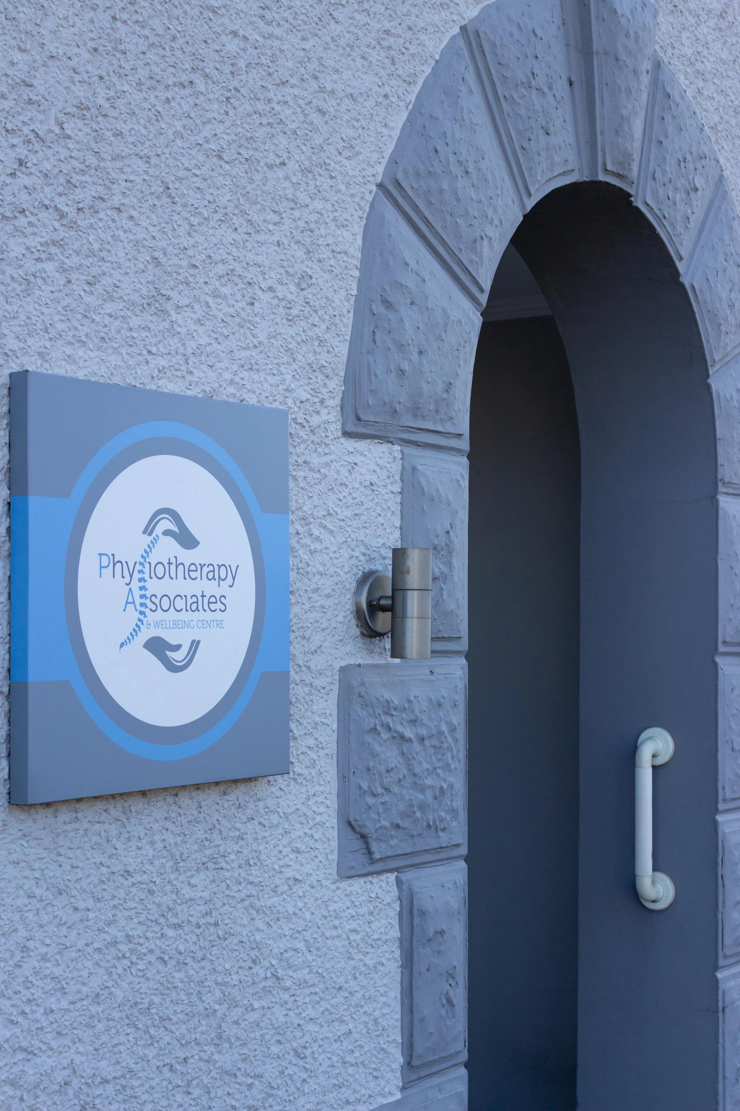 Sign for a physiotherapy and wellness center next to an arched doorway with textured wall and a white handrail.
