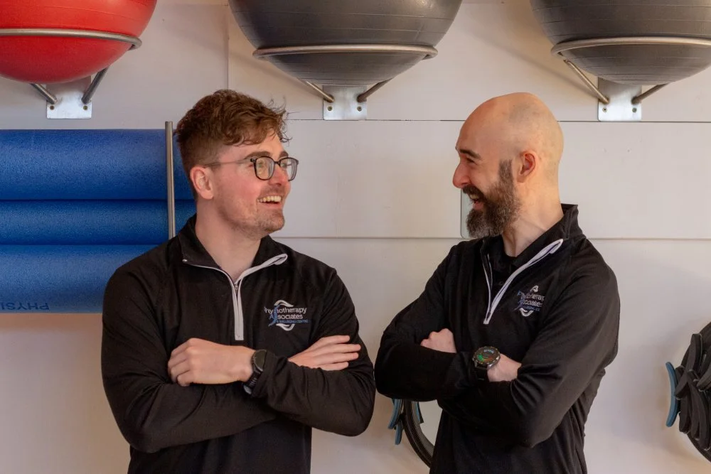 Two men in black athletic jackets smiling and talking to each other in an indoor gym with wall-mounted foam rollers and exercise balls.