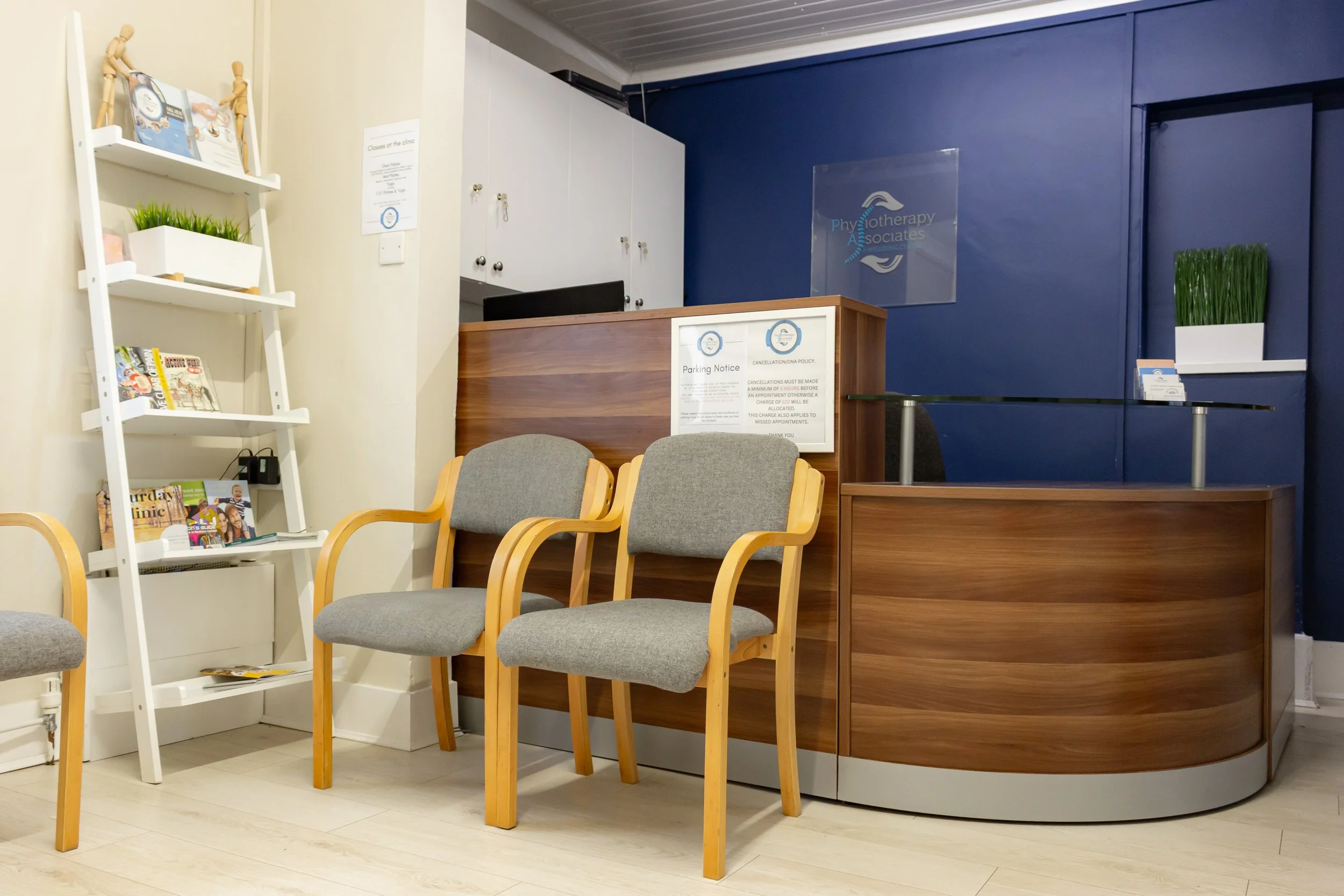 Reception area with a small desk, three gray cushioned chairs with wooden armrests, a white bookshelf with magazines, a blue wall behind the desk with a sign that reads 'Physiotherapy Associates,' and a vacuum-sealed sign on the wall.