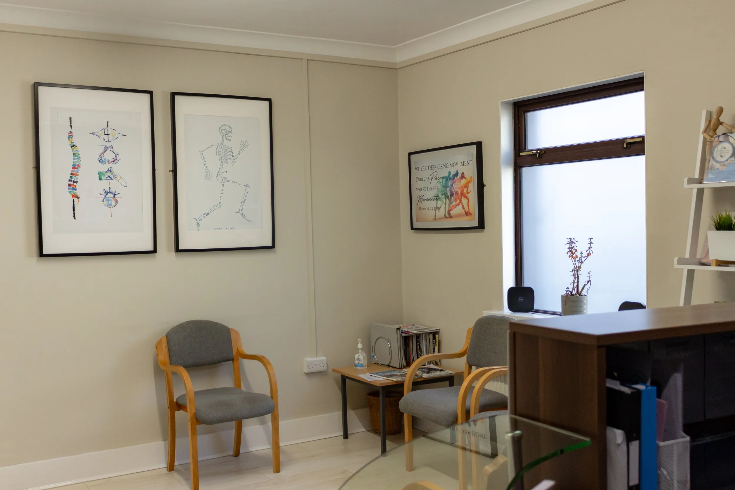 A waiting room with two gray chairs with wooden arms, a small table with magazines and a hand sanitizer, a window with a potted plant on the windowsill, framed artwork and quotes on the walls, and a bookshelf with decorative items.
