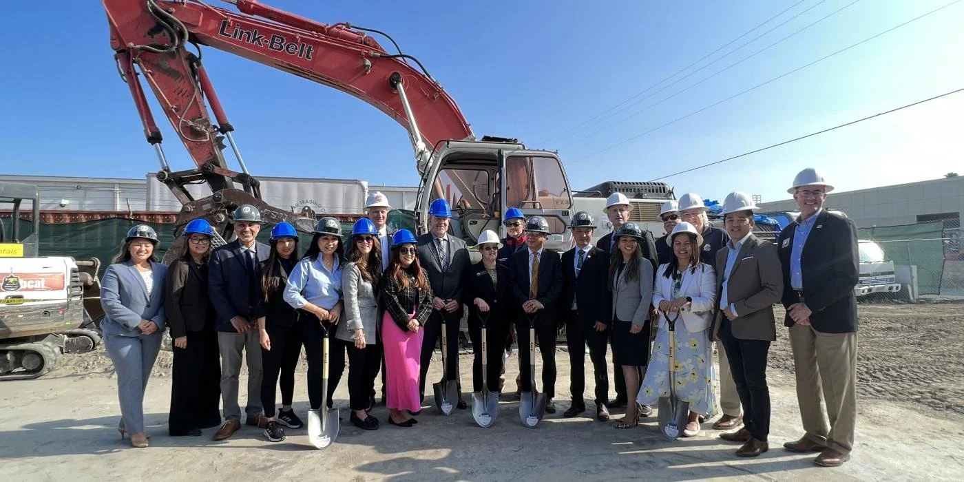 Twenty people standing in a construction site wearing hardhats and holding shovels. Behind them is large construction equipment.