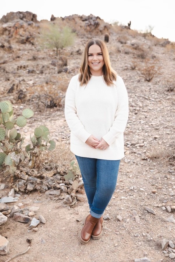A woman standing in a desert landscape with cacti and rocky terrain, wearing a white sweater, blue jeans, and brown boots, smiling at the camera.