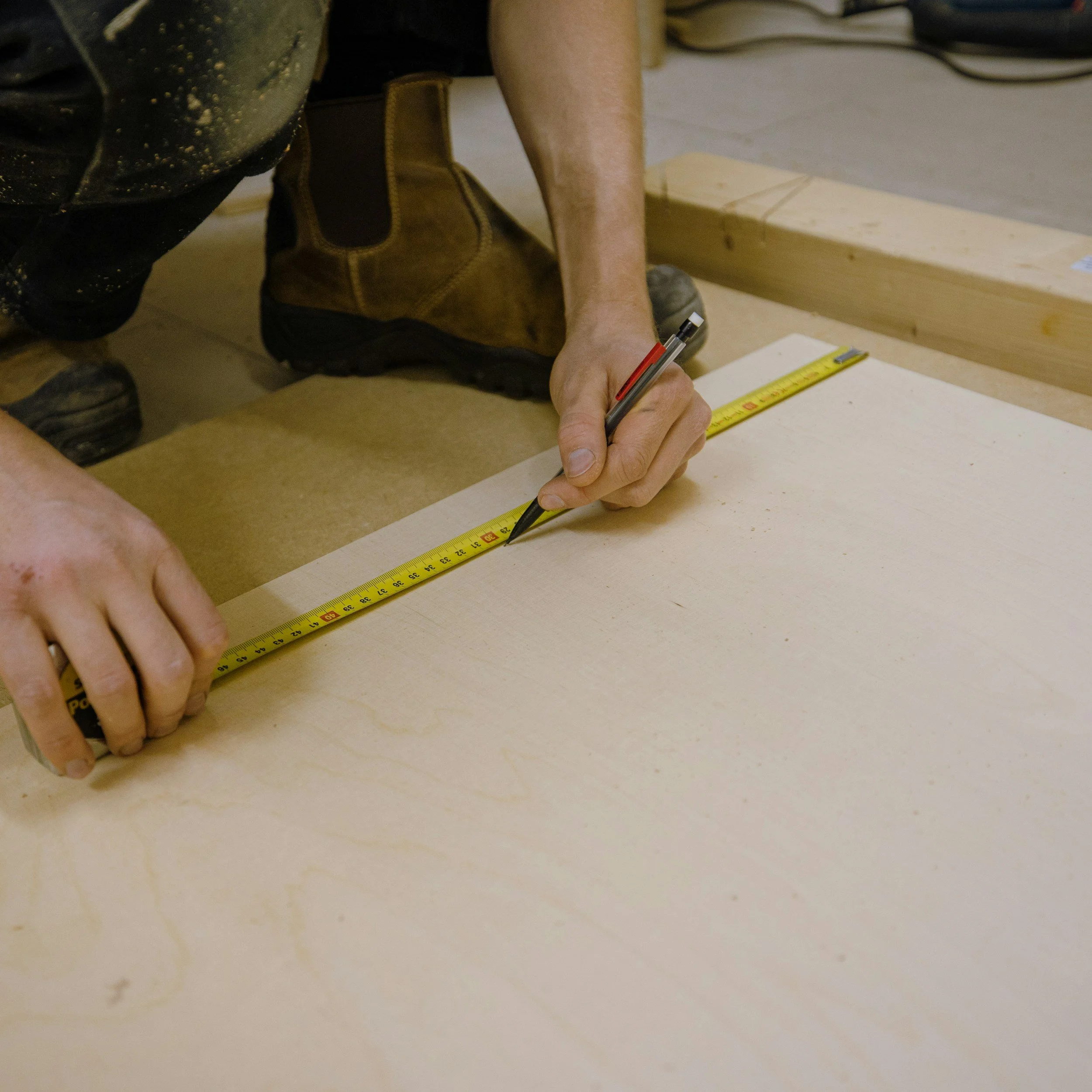 Person using a measuring tape and a pen to mark measurements on a piece of wood in a woodworking shop.