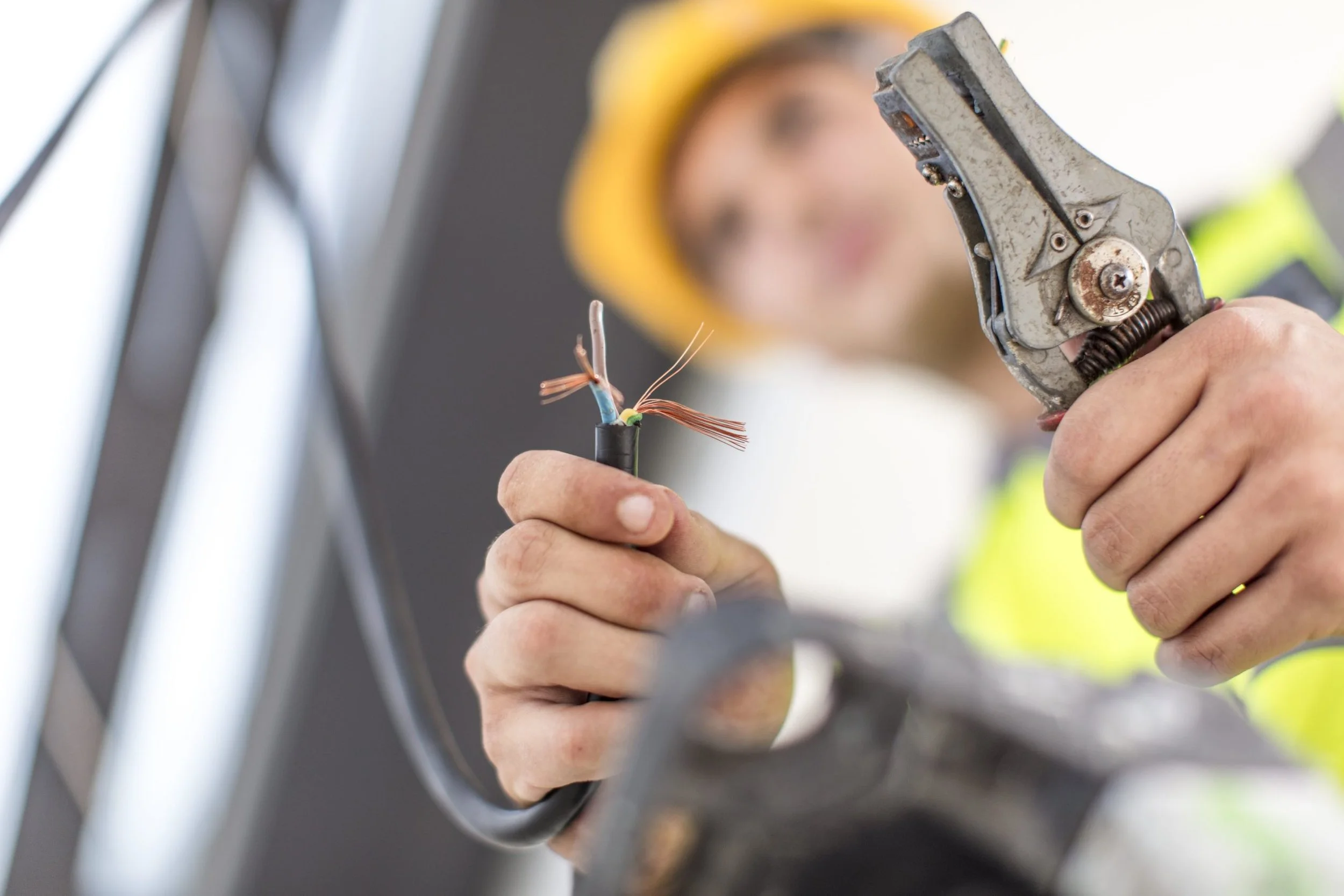 Close-up of a worker's hands holding a stripped electrical cable and using crimping tool, with a woman wearing a yellow safety helmet blurred in the background.