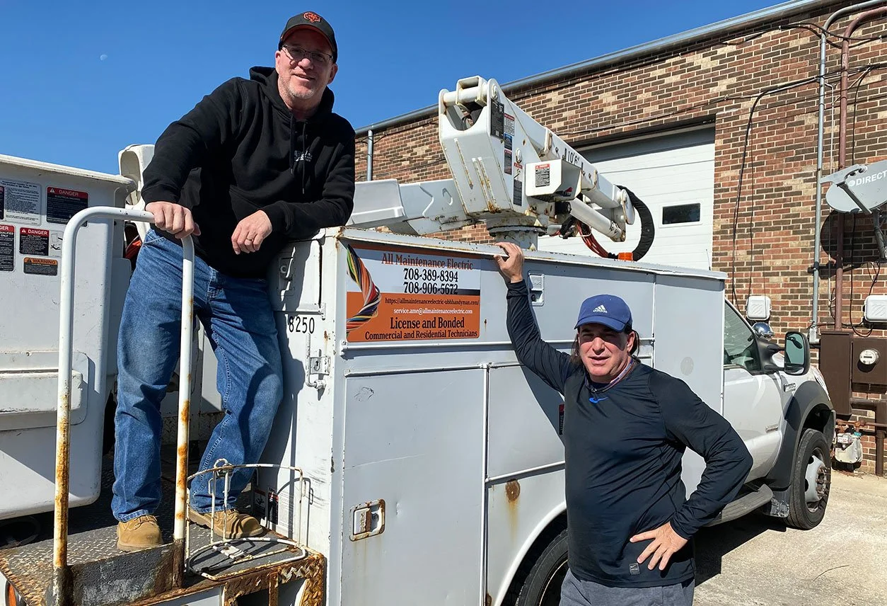 Two men standing on a work truck equipped with a bucket lift, posing for a photo outside a brick building. One is standing on the truck's platform, wearing a black hoodie and a baseball cap, while the other is standing on the ground, wearing a blue cap and black jacket. The truck has a sign with contact information and a logo for an electrical maintenance company.