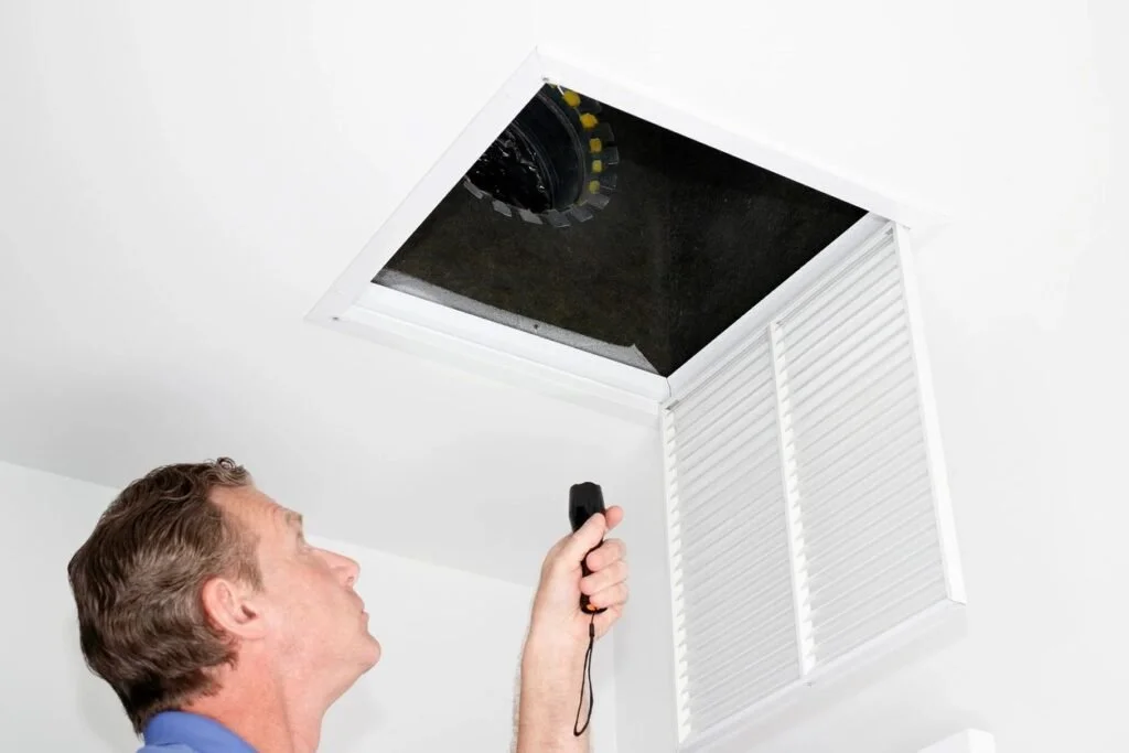 Man using a flashlight to look into an attic access hatch in a ceiling.