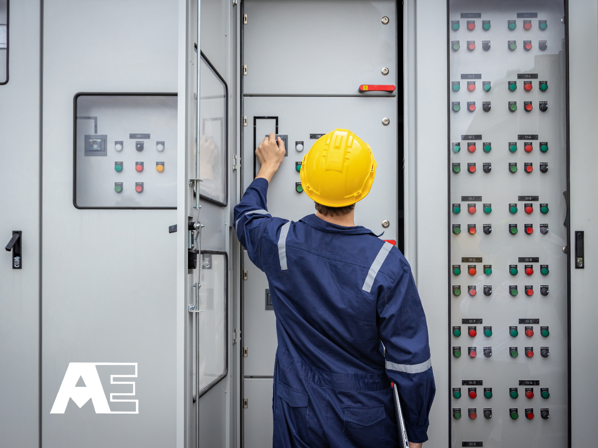A worker in a yellow safety helmet and blue uniform operating electrical control panels inside an industrial electrical cabinet.