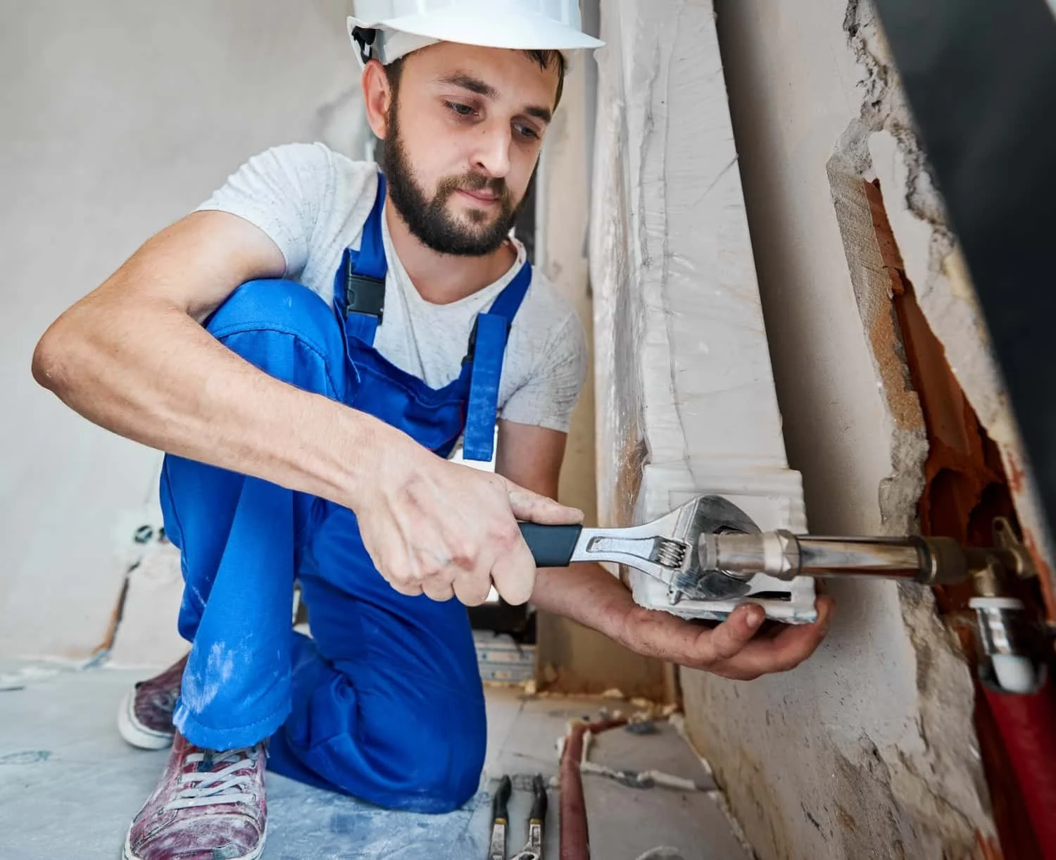 A man wearing a white helmet, gray t-shirt, and blue work overalls is kneeling and installing or repairing piping or wall fixtures with an adjustable wrench in a building under construction or renovation.