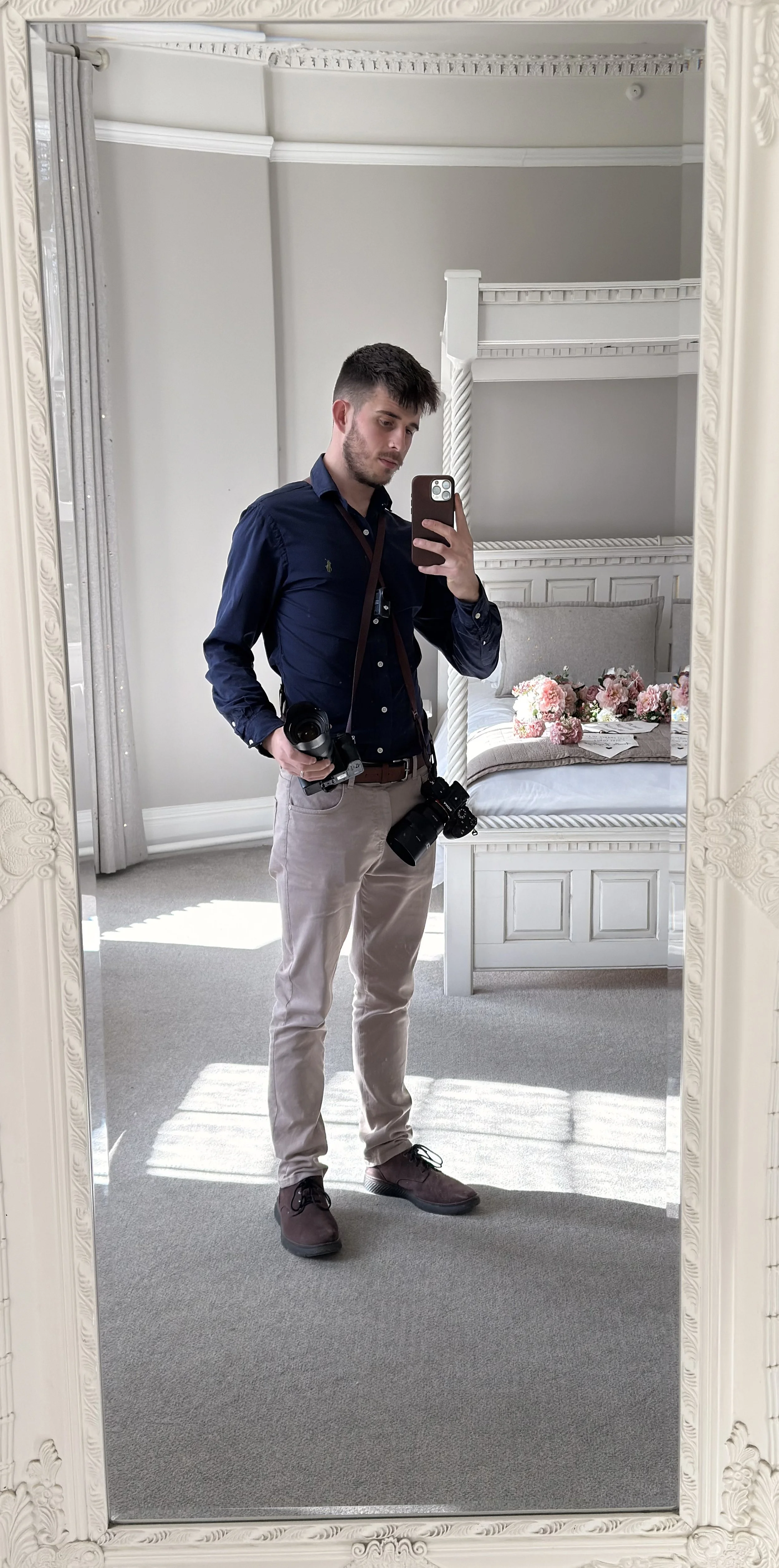 A man taking a selfie in a mirror in a well-decorated room with a bed, floral bedding, and curtains. He is holding two cameras and dressed in a navy shirt and light-colored pants.