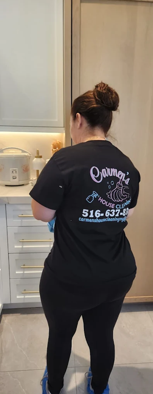 Carmen's House Cleaning LLC's staff member with her hair in a bun standing in a kitchen, facing a counter and shelves. She is wearing a black T-shirt with the text 'Carminda's House Cleaning' on the back and glasses of cleaning supplies are nearby.