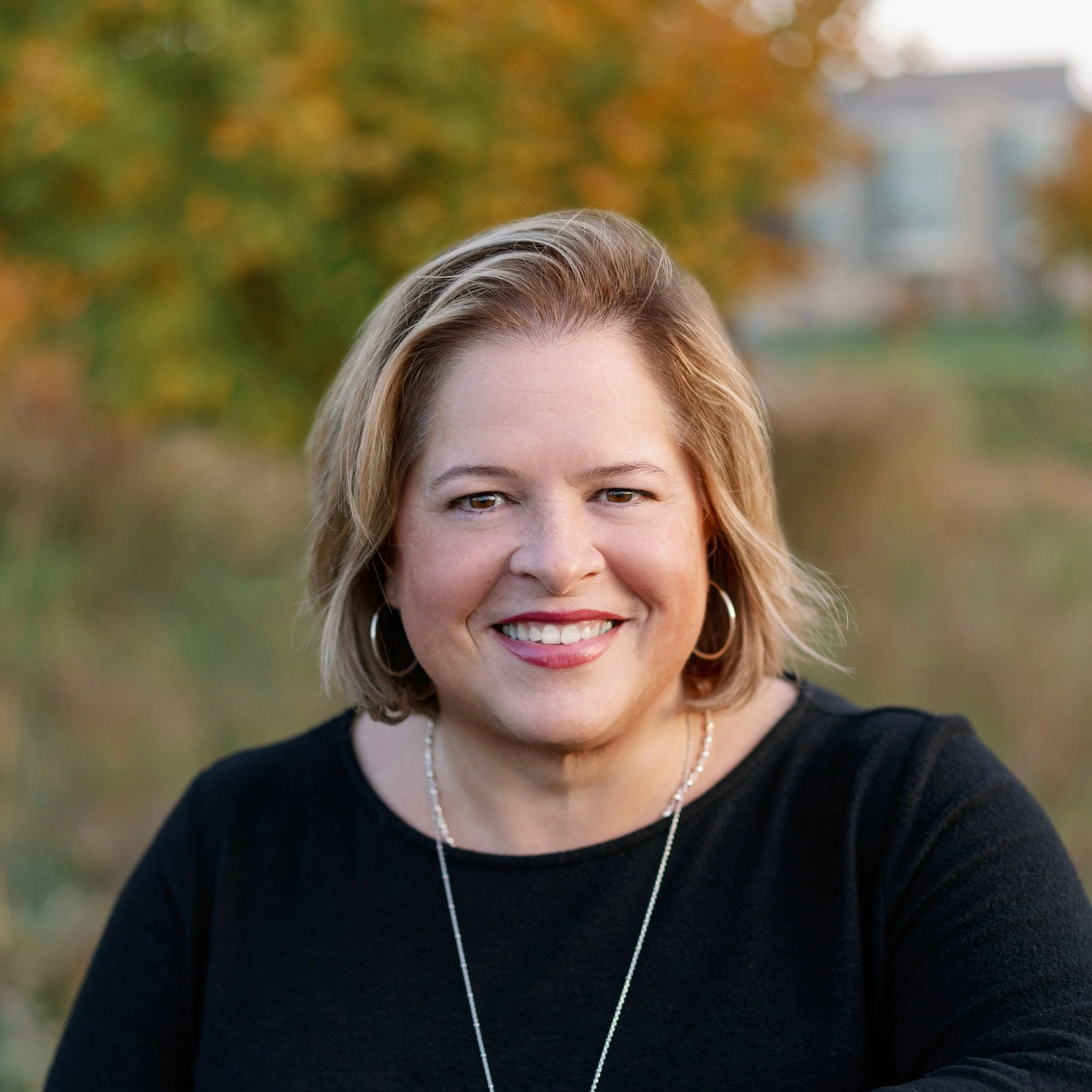 Eva Buck smiling outdoors with autumn trees in the background.