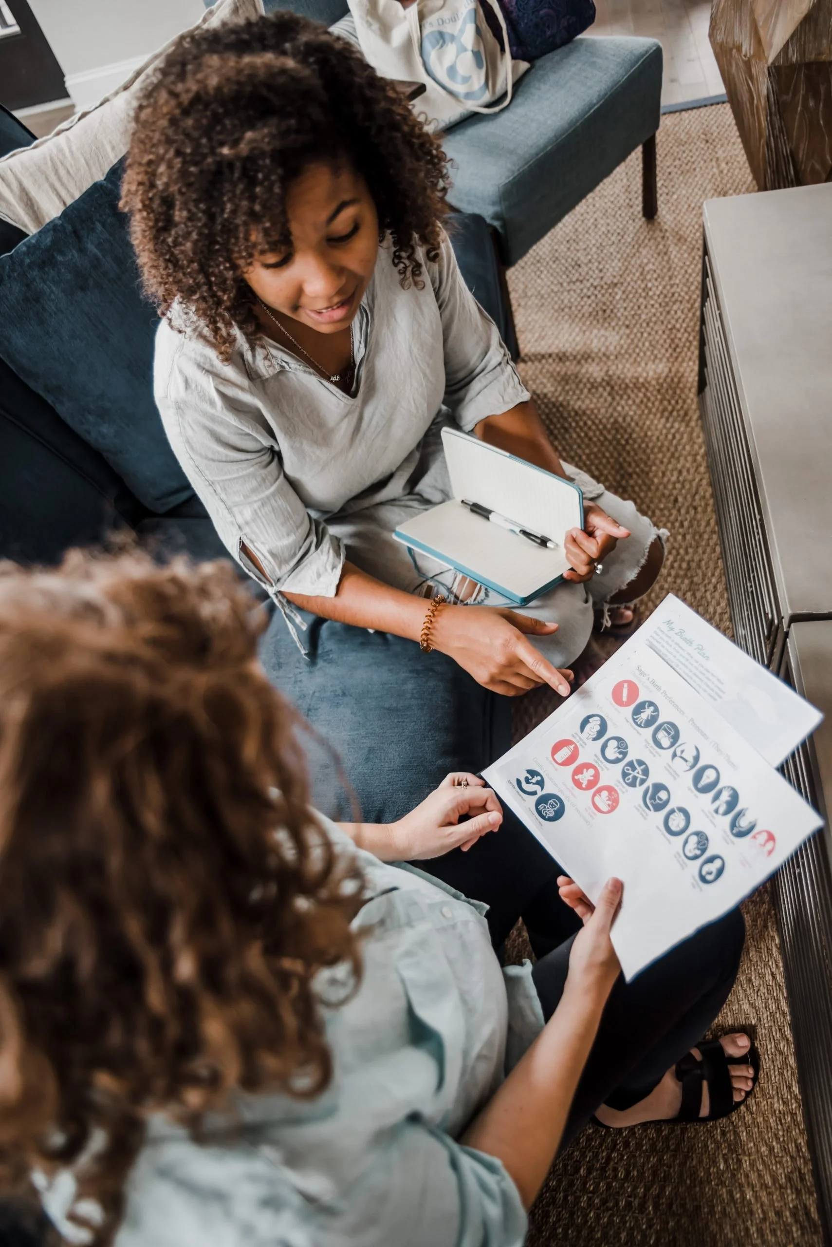 A group of women sitting on a carpeted floor, engaged in a discussion or presentation. One woman is holding a printed infographic with icons and text. Another woman is taking notes in a small notebook with a pen. There are tables and couches in the background.