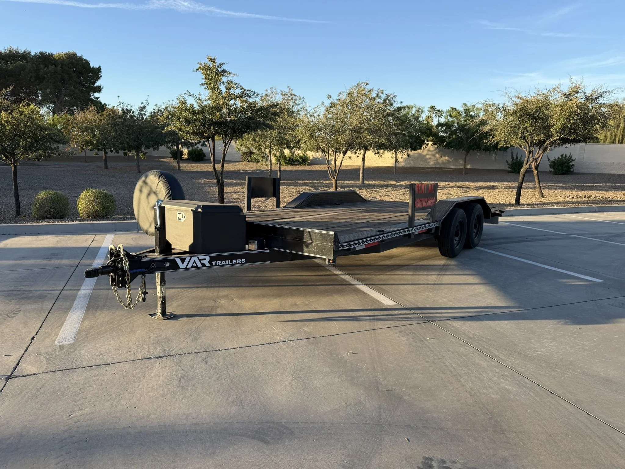 Empty parking lot with a flatbed trailer and a sign that reads "Rent Me"; background includes small trees, shrubs, and a clear blue sky.