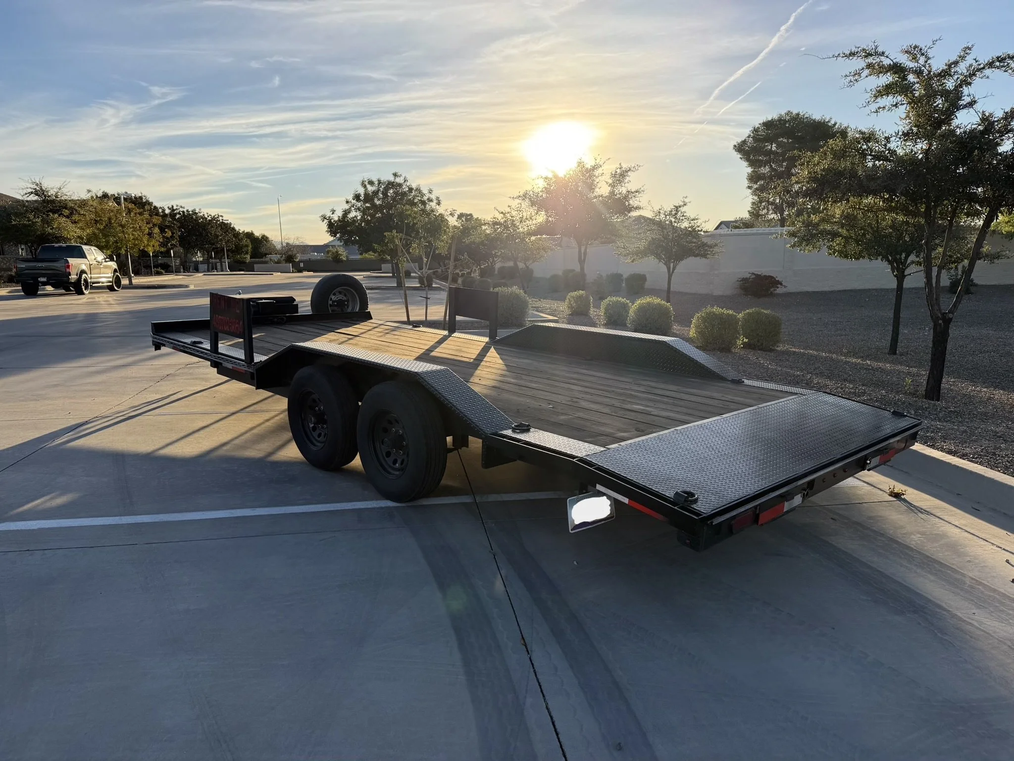 A flatbed trailer parked on a parking lot during sunset with trees and a white wall in the background.