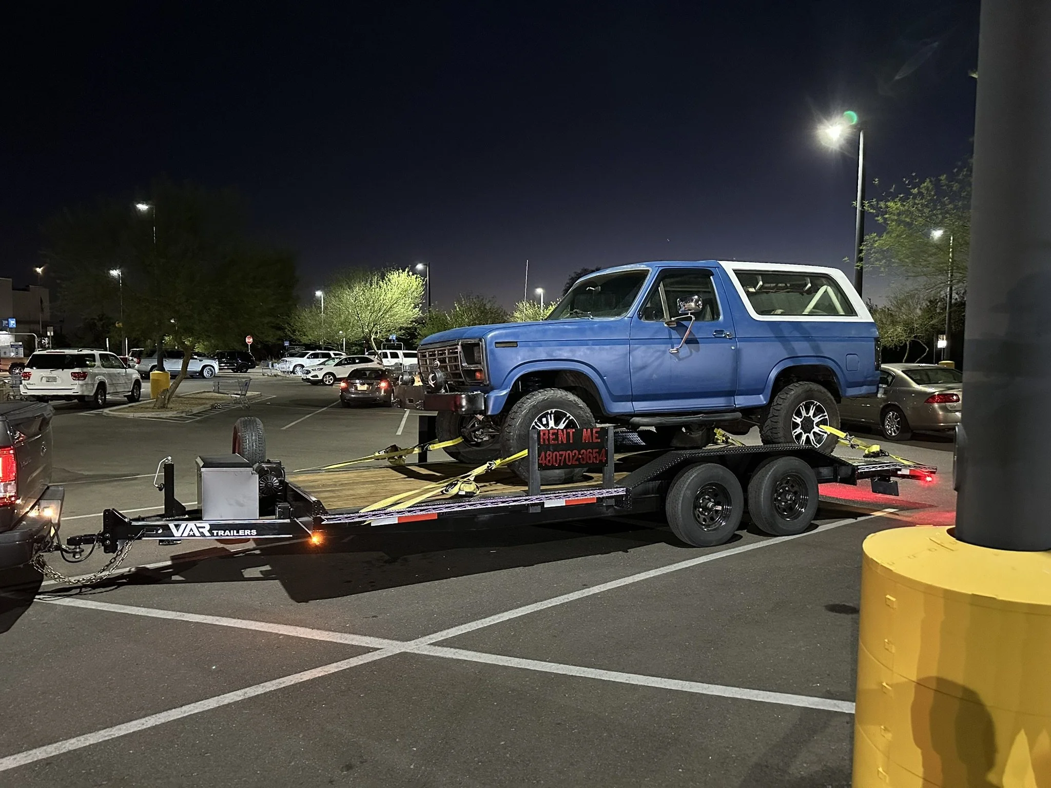 A blue vintage Ford Bronco SUV on a trailer in a parking lot at night. The trailer has a sign that says 'Rent Me' with a phone number. Surrounding parked cars and trees are visible under streetlights.