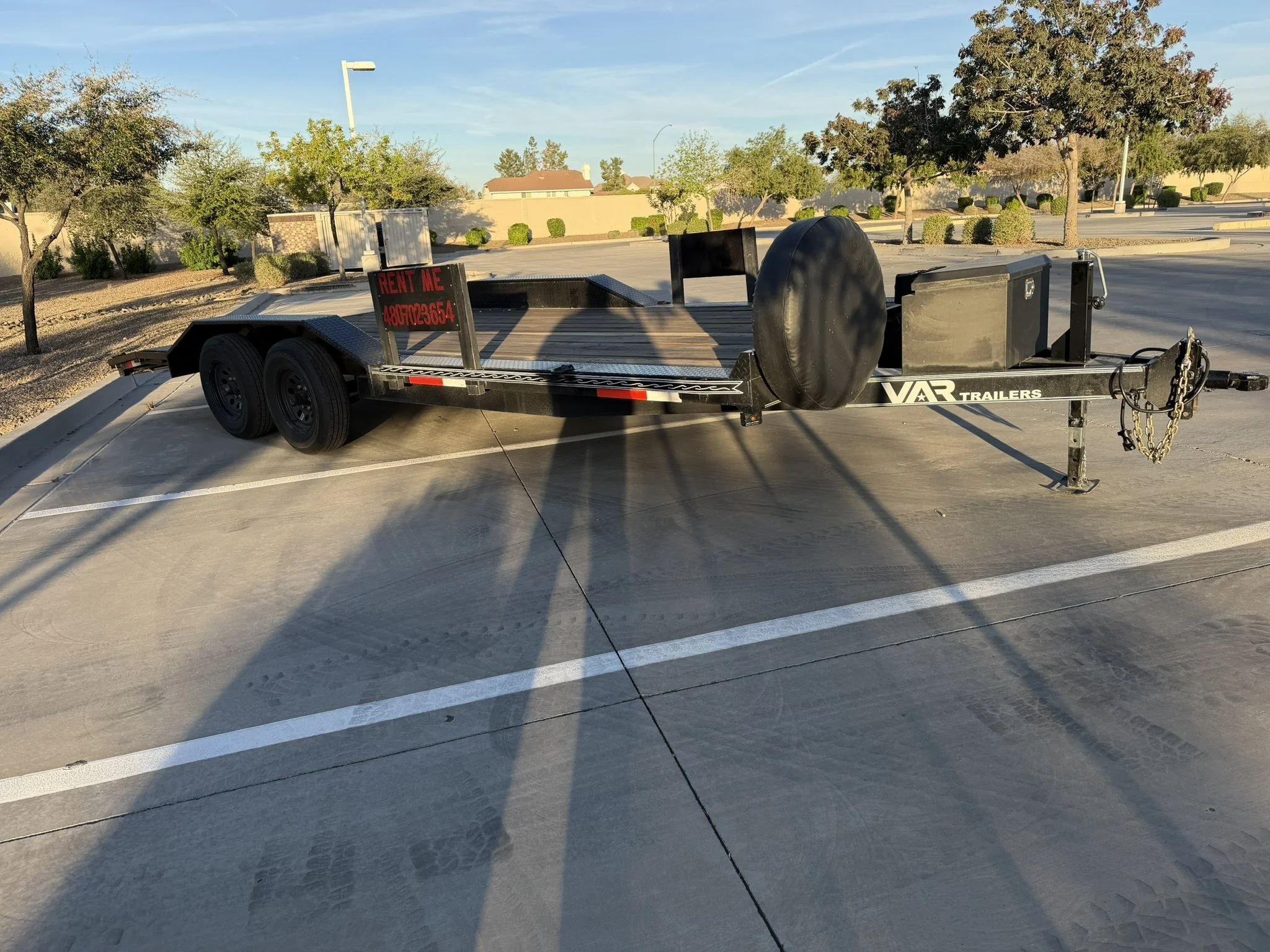 Empty parking lot with a flatbed trailer in the foreground, trees, bushes, and residential houses in the background, under a clear blue sky.
