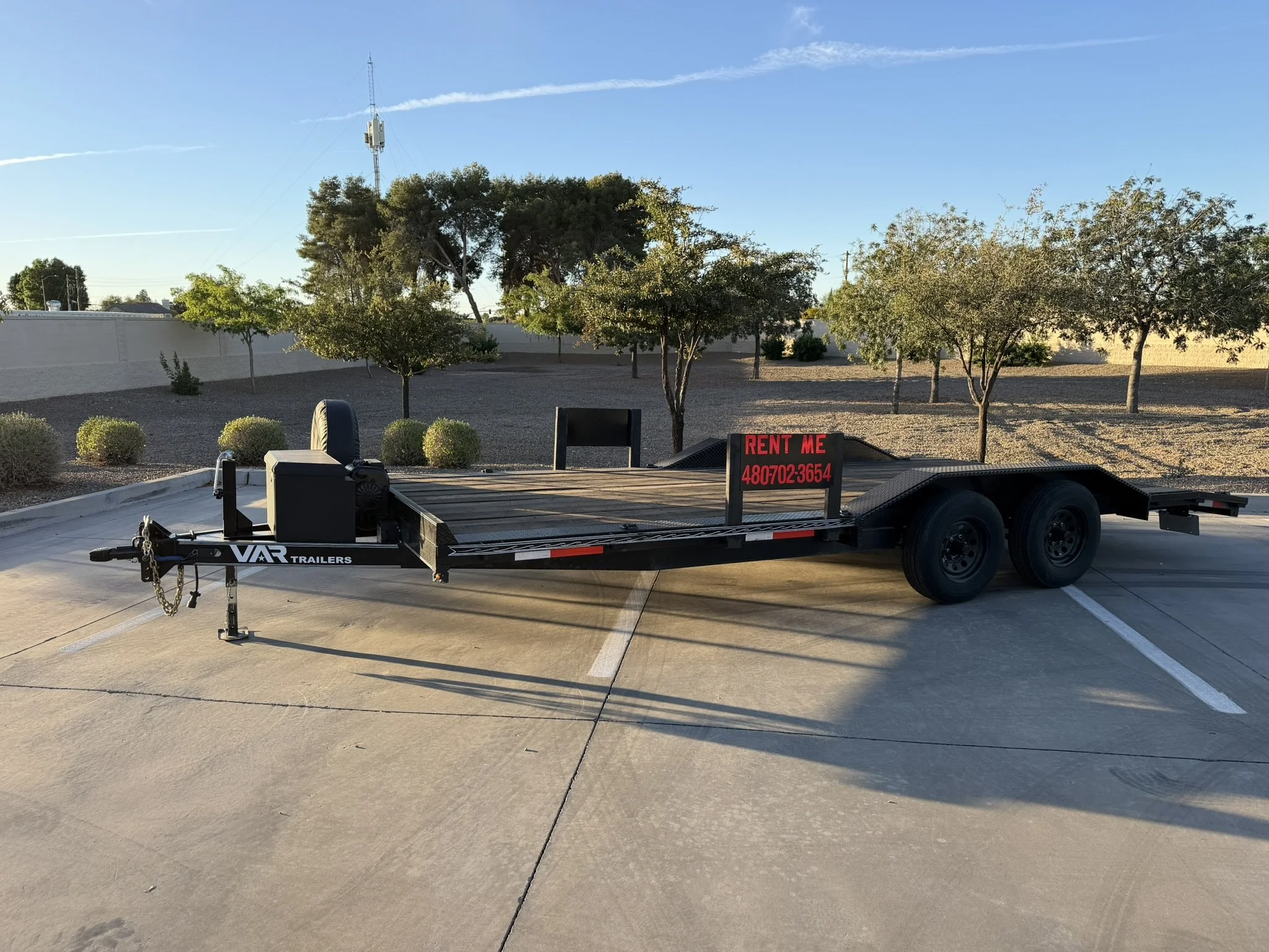 Empty parking lot with a flatbed trailer, a sign that reads 'Rent Me' with a phone number, several small trees, and a larger tree in the background under a clear blue sky.