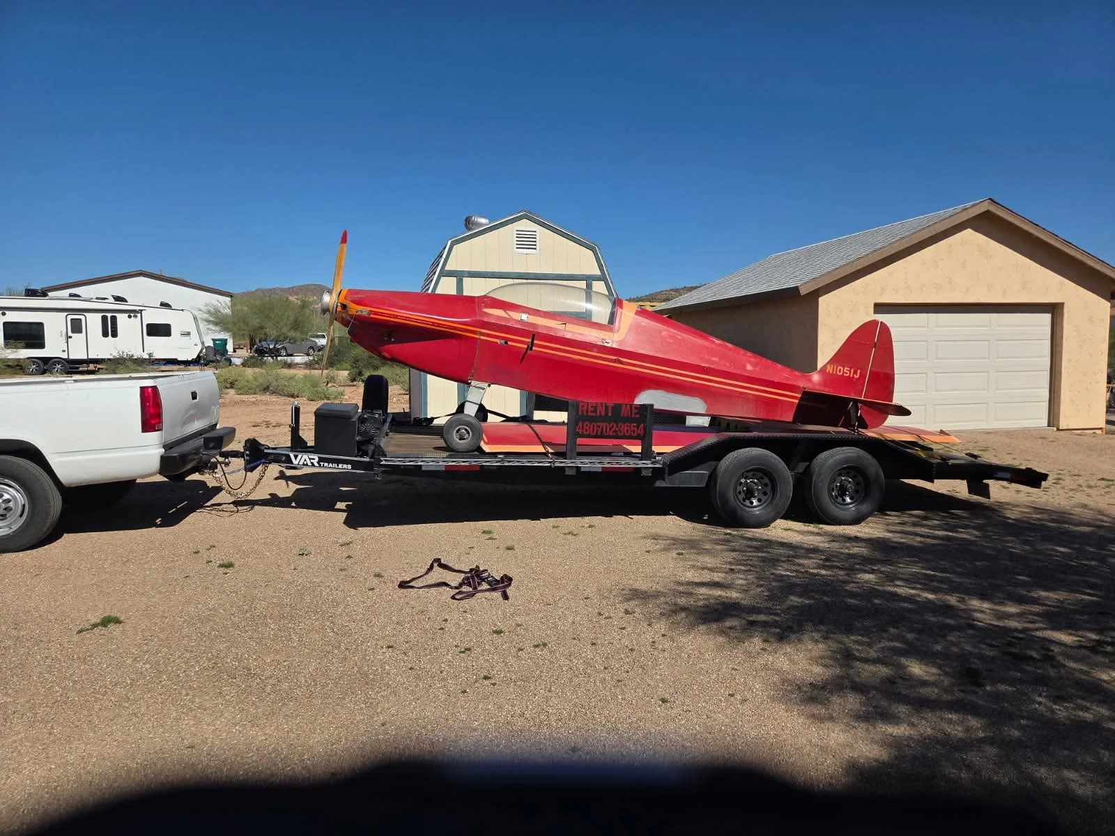 Red small airplane on a trailer hitched to a white pickup truck, parked on a gravel driveway in front of a residential garage and houses, under a clear blue sky.