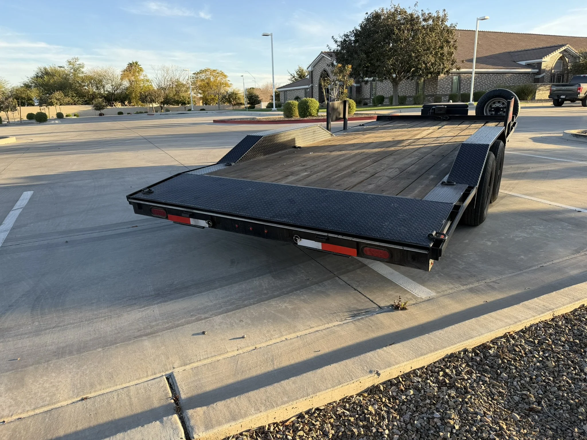 Empty parking lot with a flatbed trailer in the foreground, several trees and residential buildings in the background, and a clear blue sky.