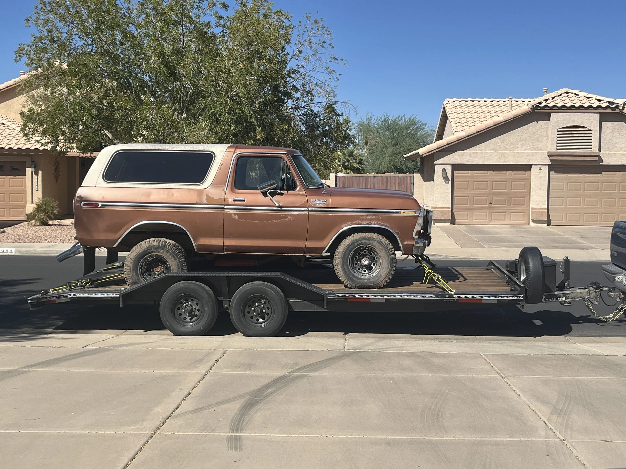 A brown vintage SUV is loaded onto a black flatbed trailer in a suburban driveway with tan houses and a large tree in the background.