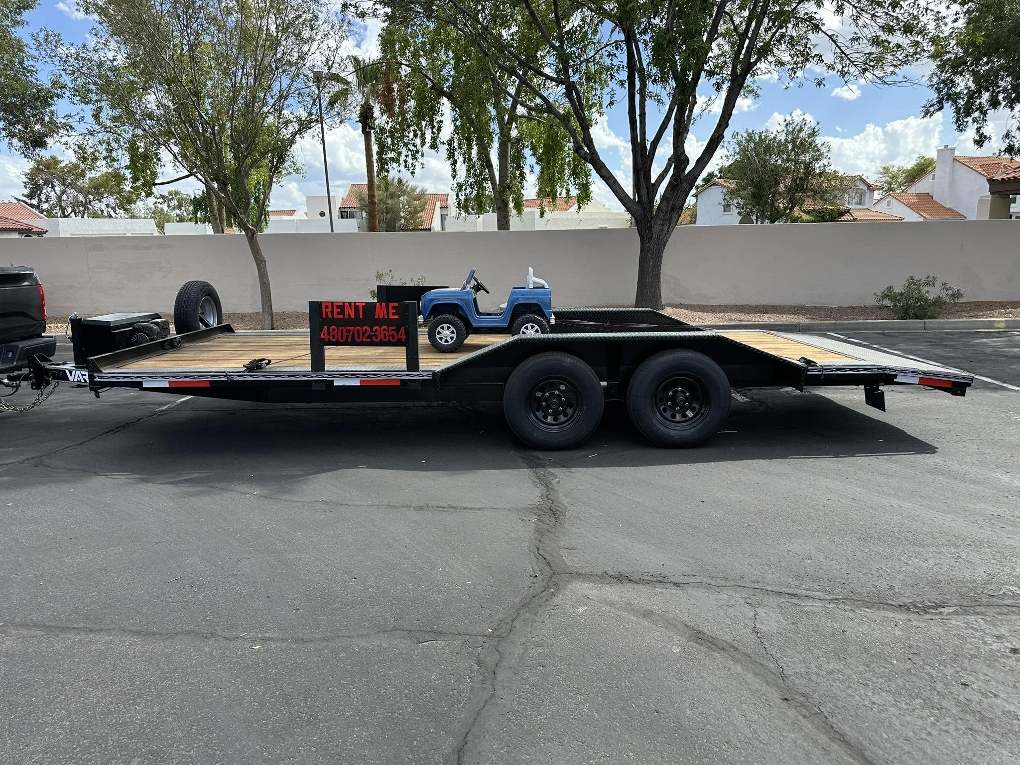 A parking lot with a black flatbed trailer carrying a small plastic blue toy car and a black sign that reads 'RENT ME' with a phone number. There are trees and residential houses in the background.