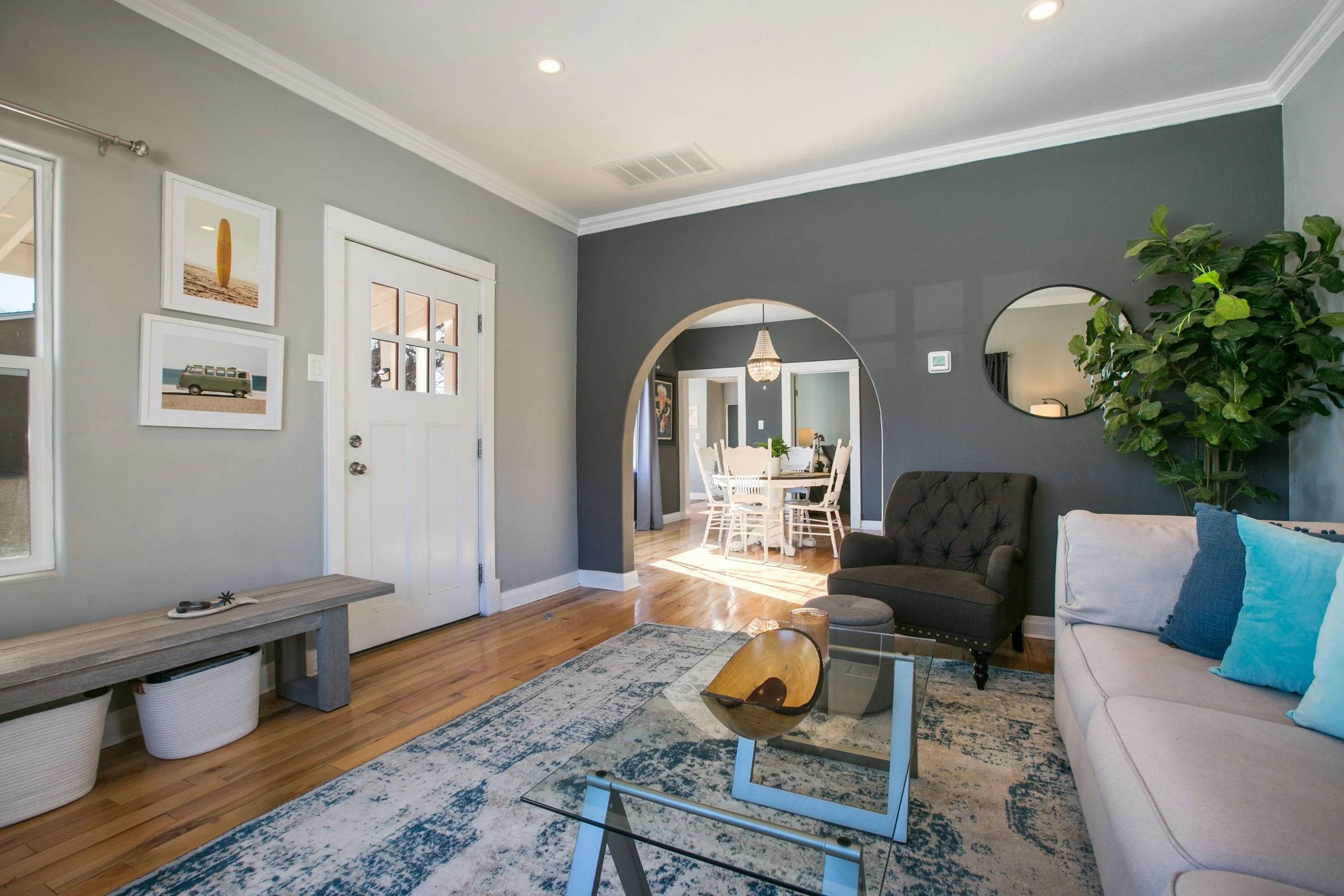 Living room view looking into dining area with a round table and white chairs, dark gray and light gray walls, wooden floors, and decor including paintings, mirror, and plants.