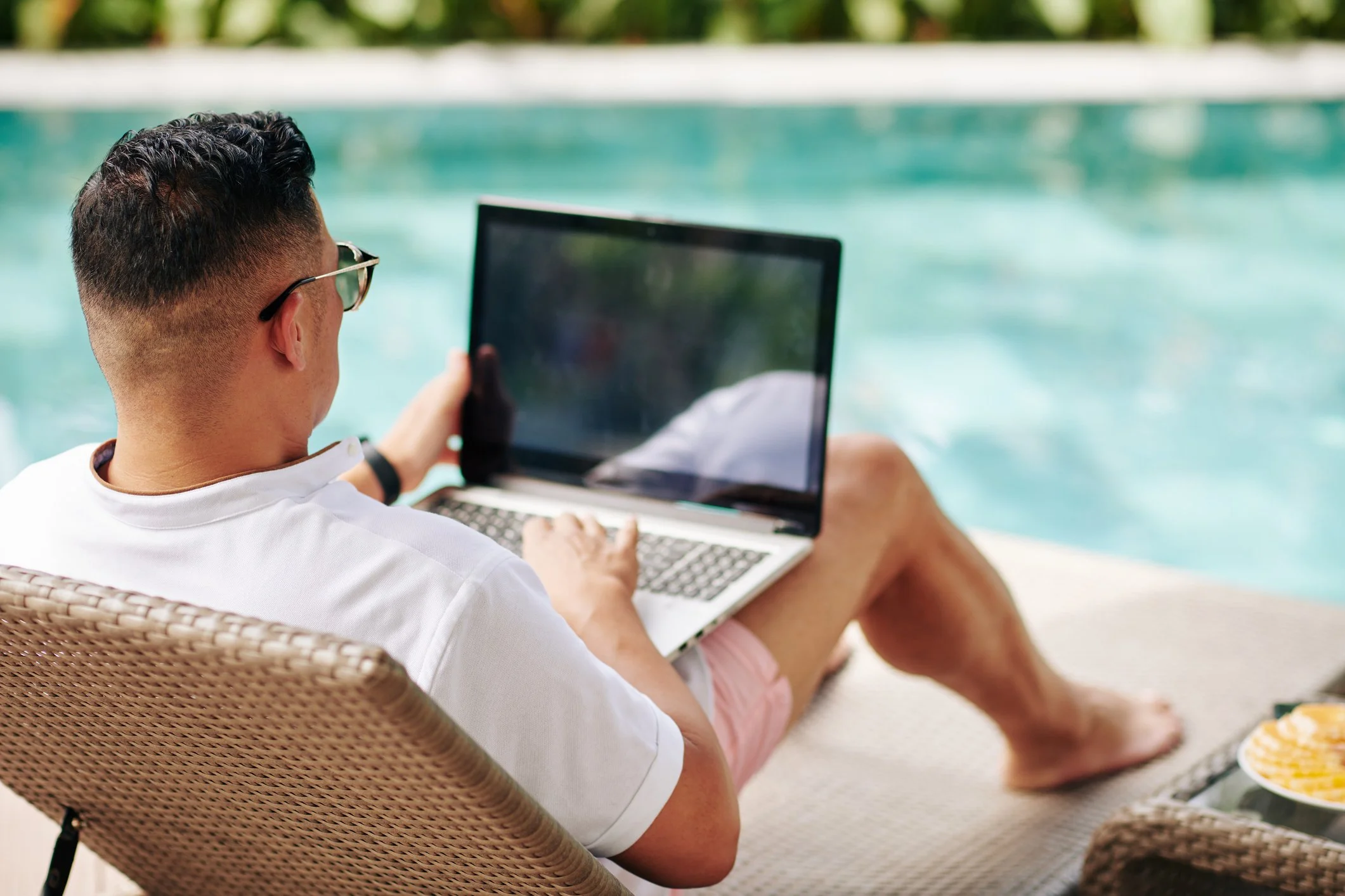 Man sitting by a pool working on a laptop, wearing sunglasses and a white shirt.