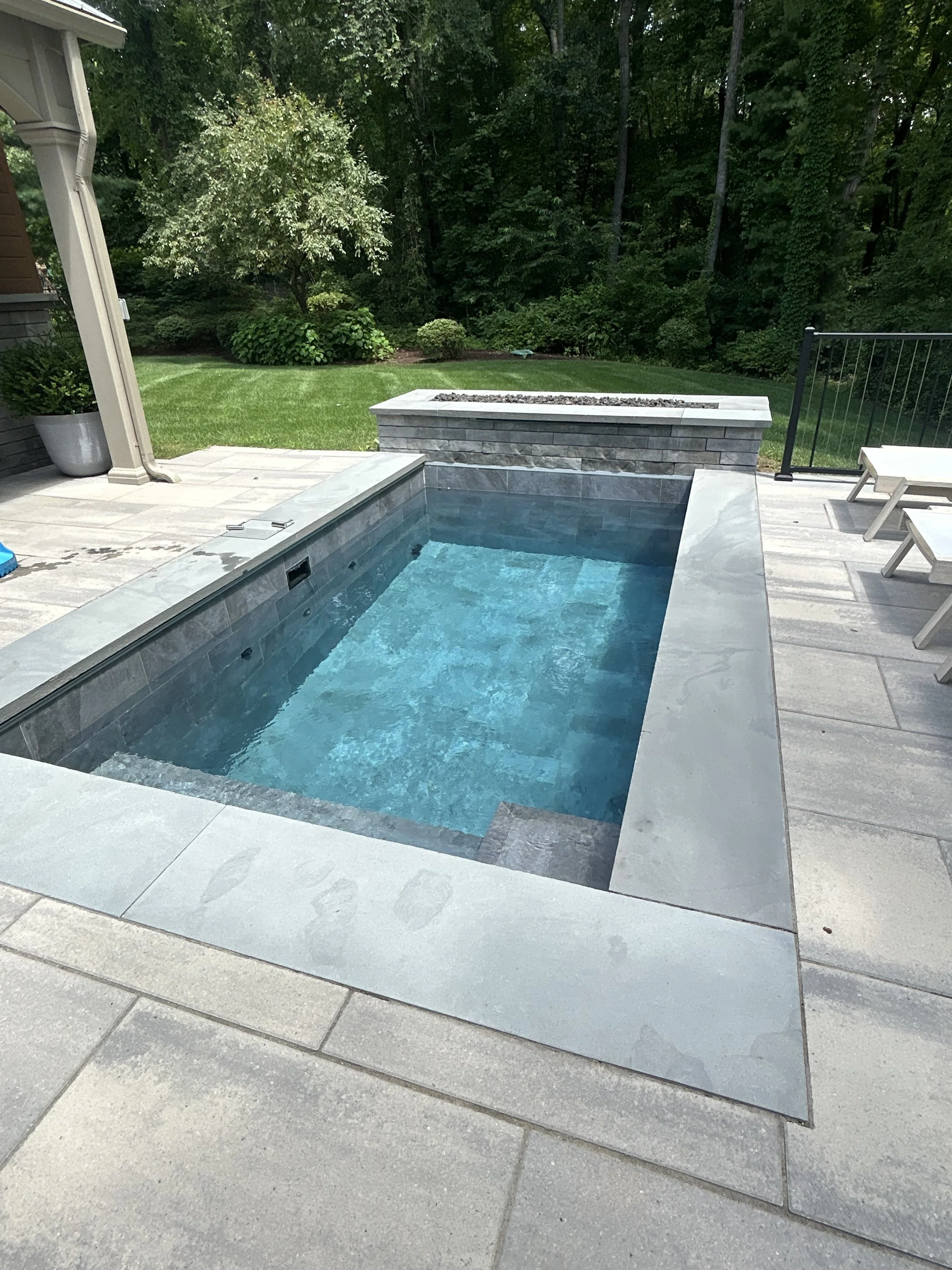 A modern rectangular in-ground swimming pool with light gray stone surround on a patio, adjacent to a grassy yard and surrounded by trees.