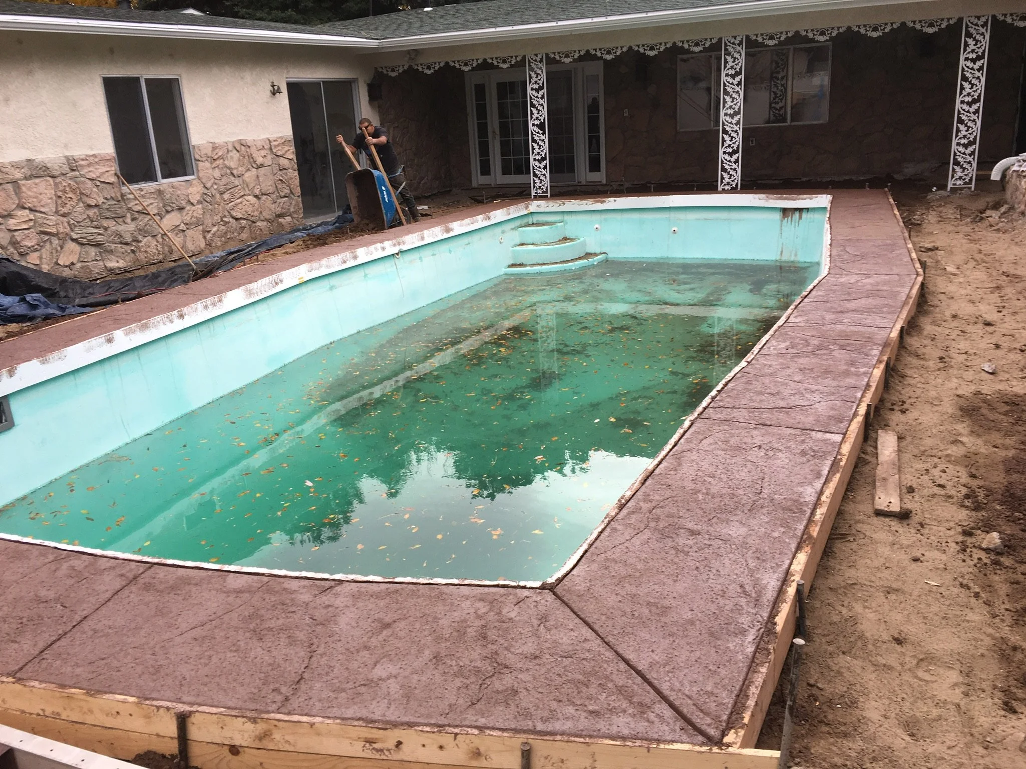 A backyard with a swimming pool under renovation, filled with dirty water and leaves, surrounded by construction materials and partially built patio.