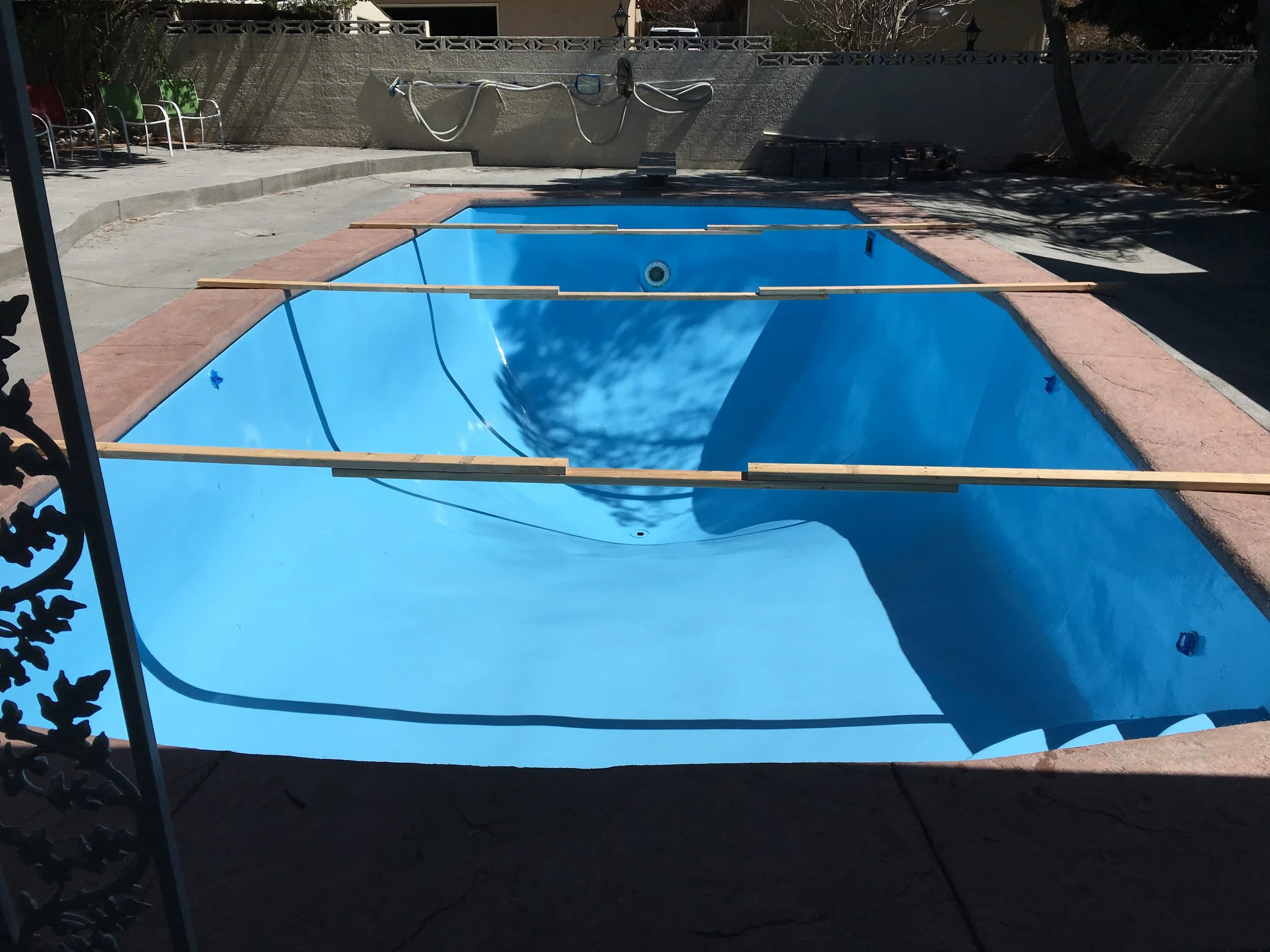 Empty swimming pool with blue interior and wooden planks across top, located outdoors in a sunny area with trees and a beige wall in the background.
