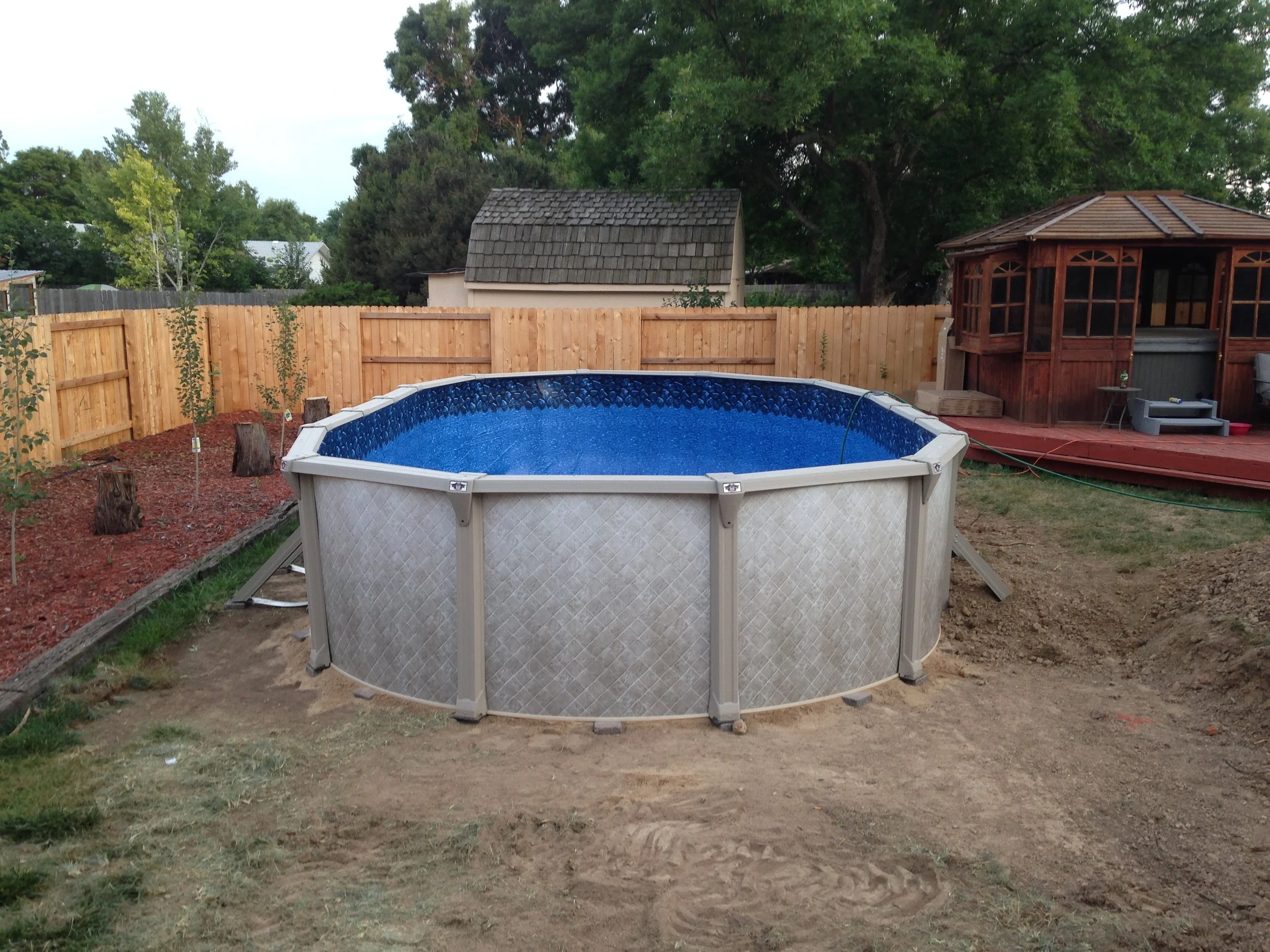 Above ground oval swimming pool with a blue interior in a backyard, surrounded by a wooden privacy fence, with trees, small structures, and a garden area nearby.
