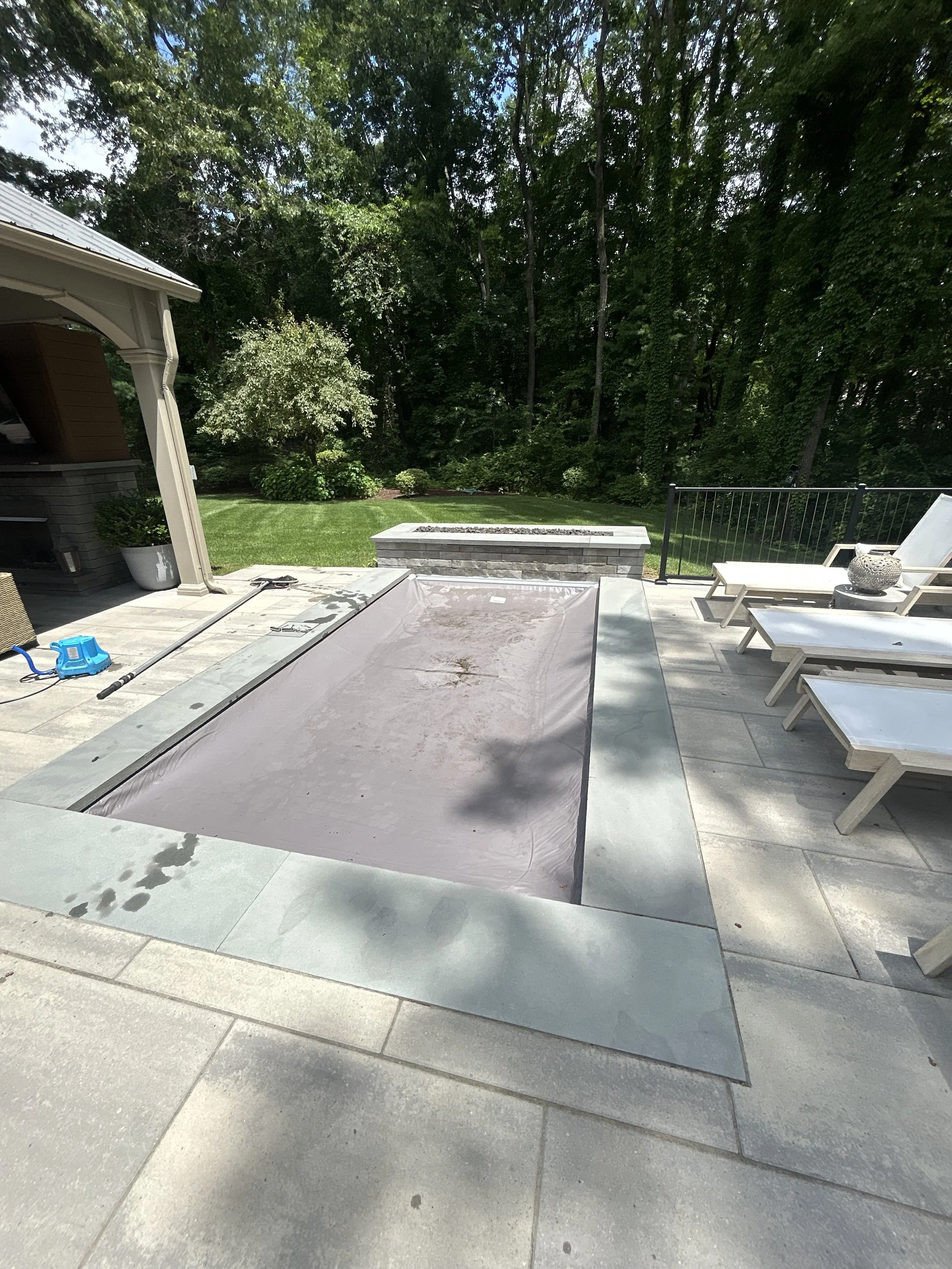 Empty hot tub covered with a beige tarp on a patio with lounge chairs and lush green trees in the background.