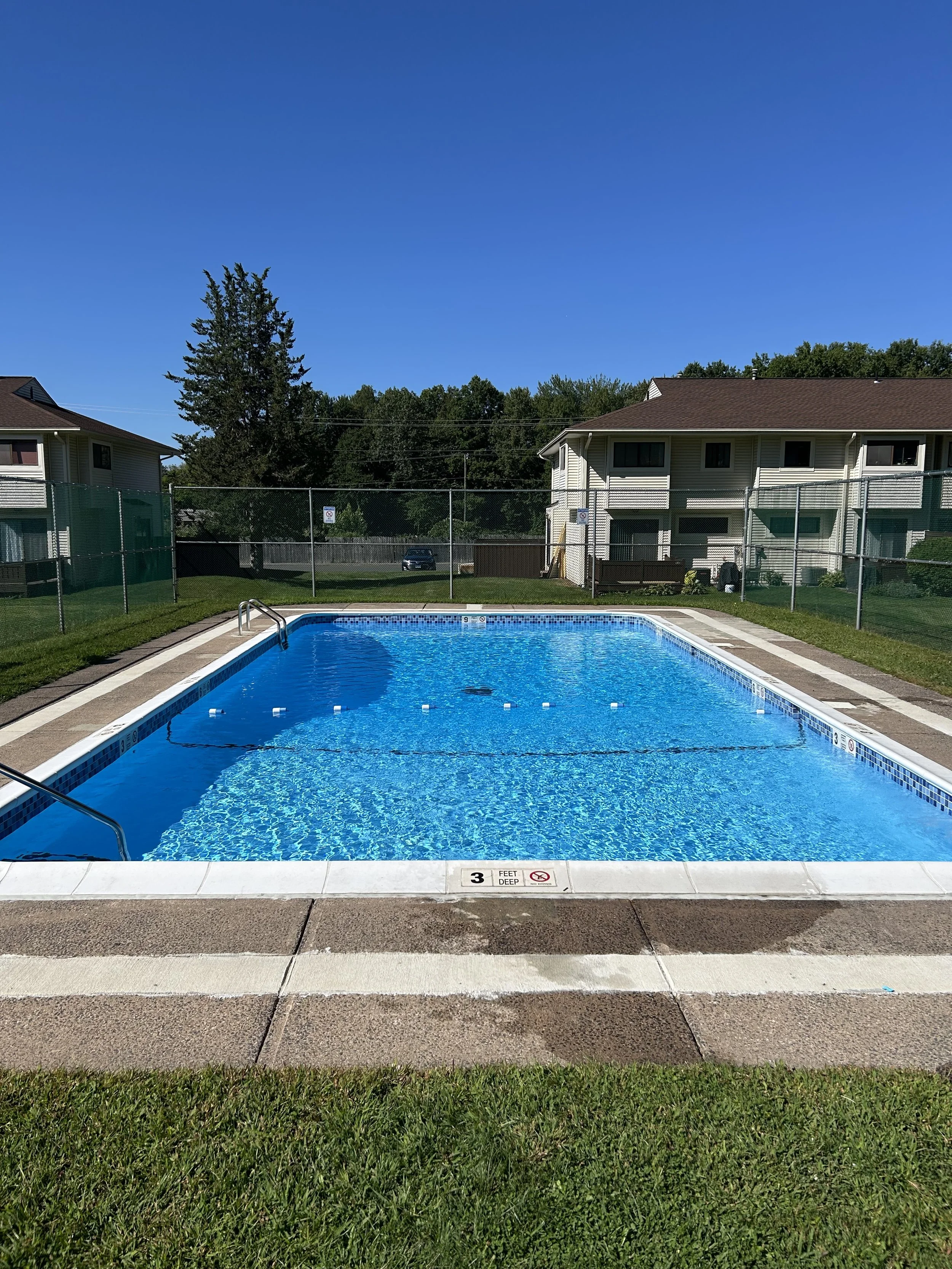 Outdoor swimming pool surrounded by a fence, with apartment buildings in the background and a clear blue sky.