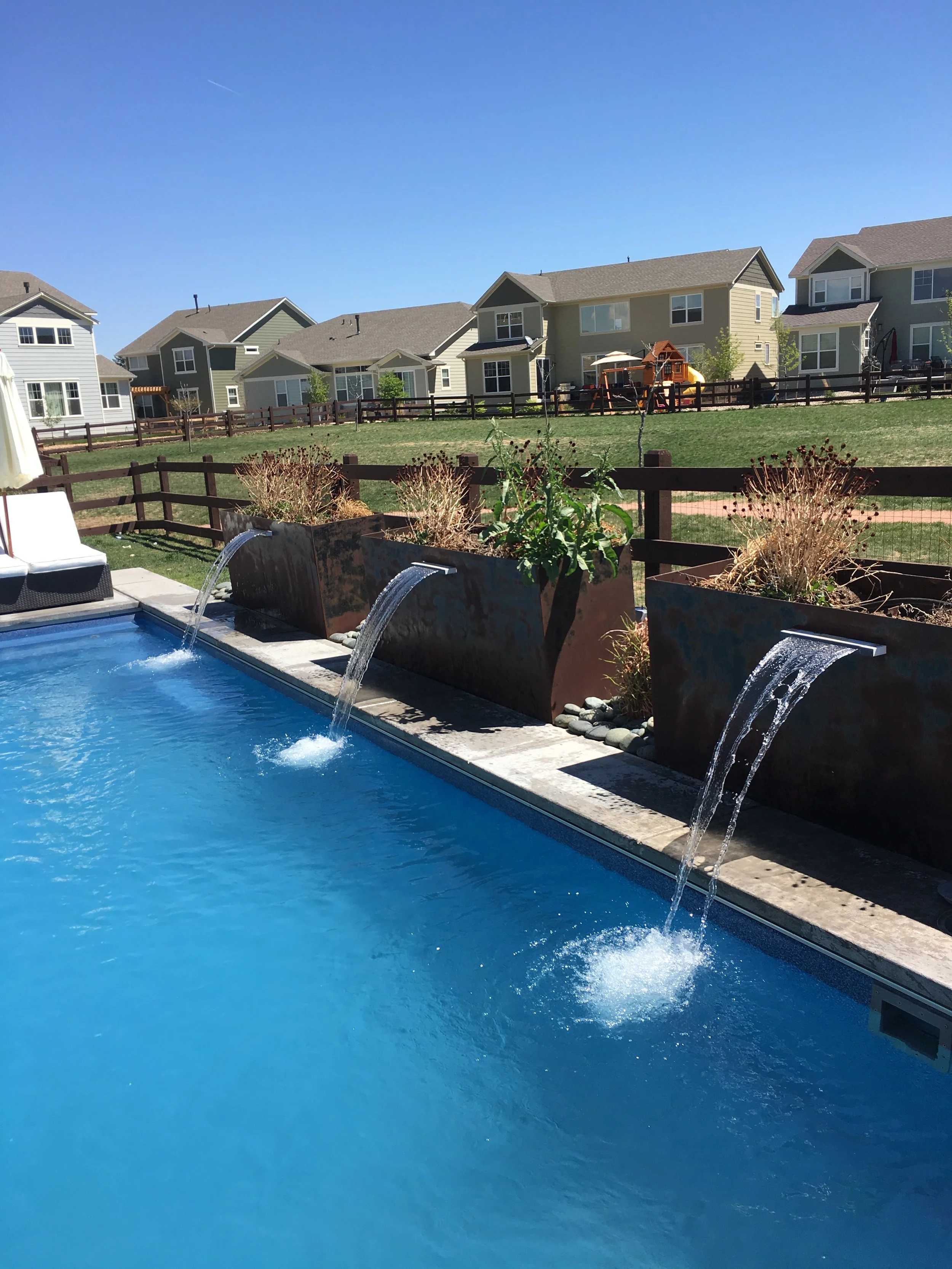 A backyard swimming pool with three water features and a row of metal planters with plants, set against a grassy yard and several houses under a clear blue sky.