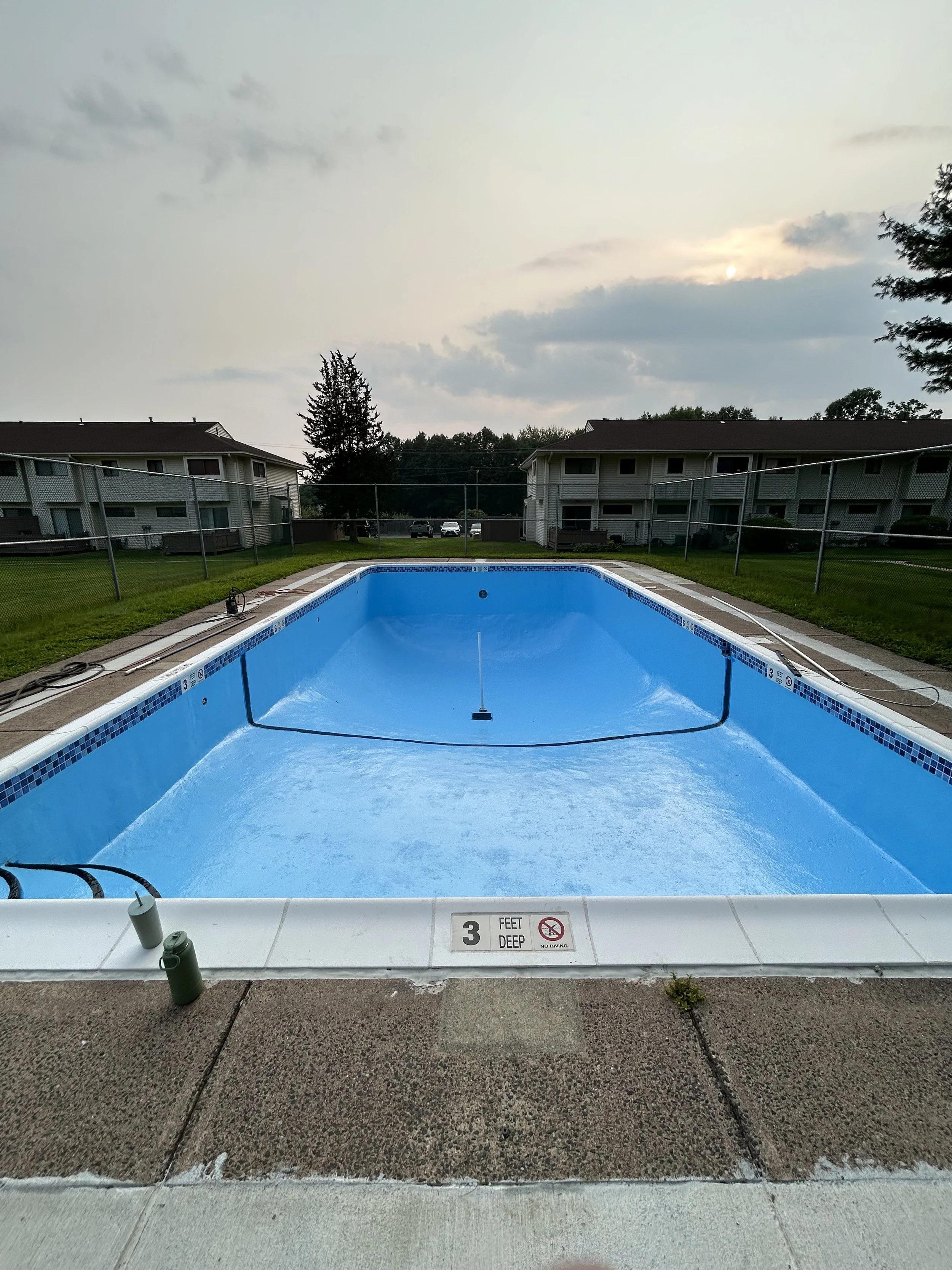 Empty swimming pool with a depth of 3 feet, surrounded by a concrete deck, with a safety chain, and buildings in the background under a cloudy sky at sunset.